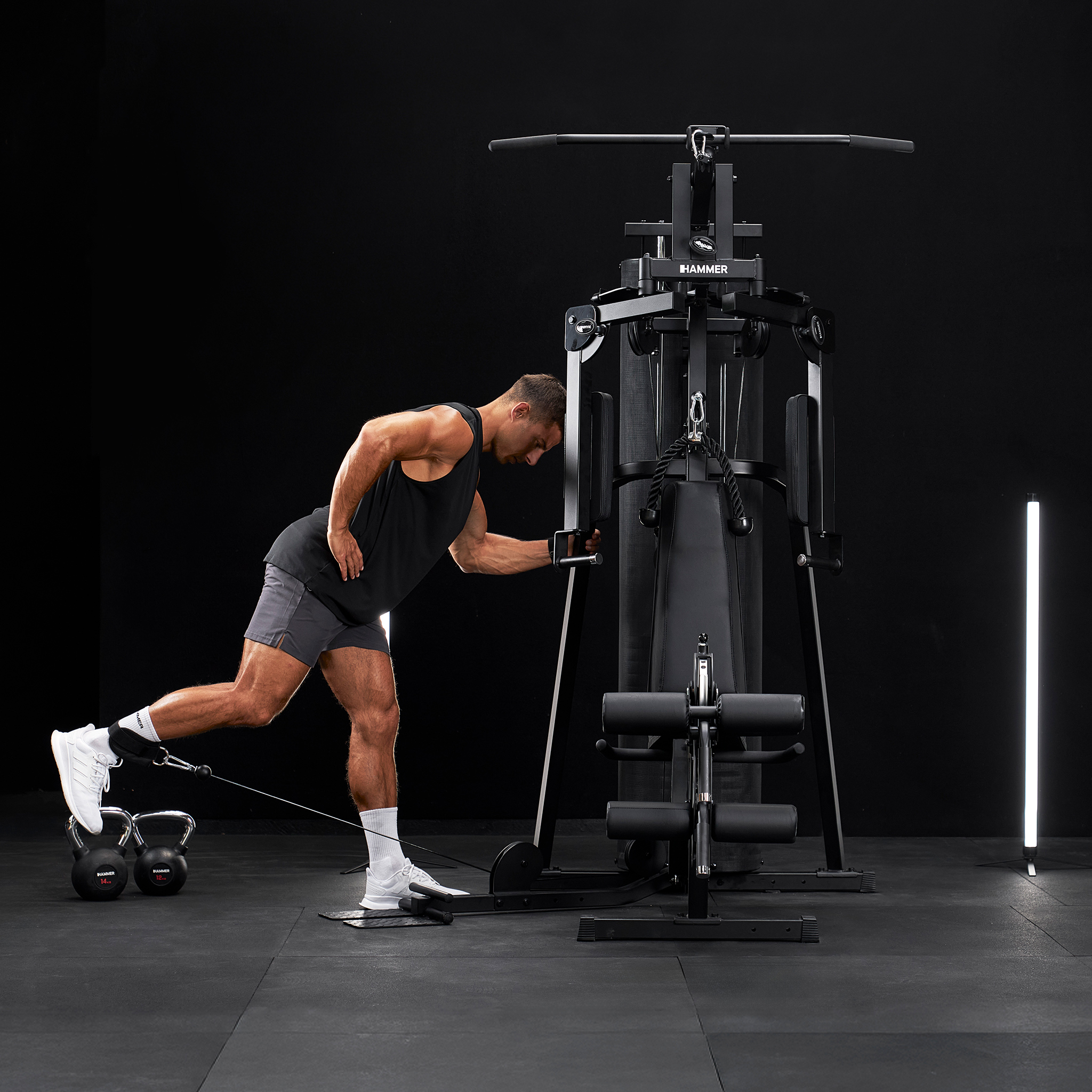 Mann trainiert Bein mit Kabelzug an Fitnessgerät, Kettlebells daneben.