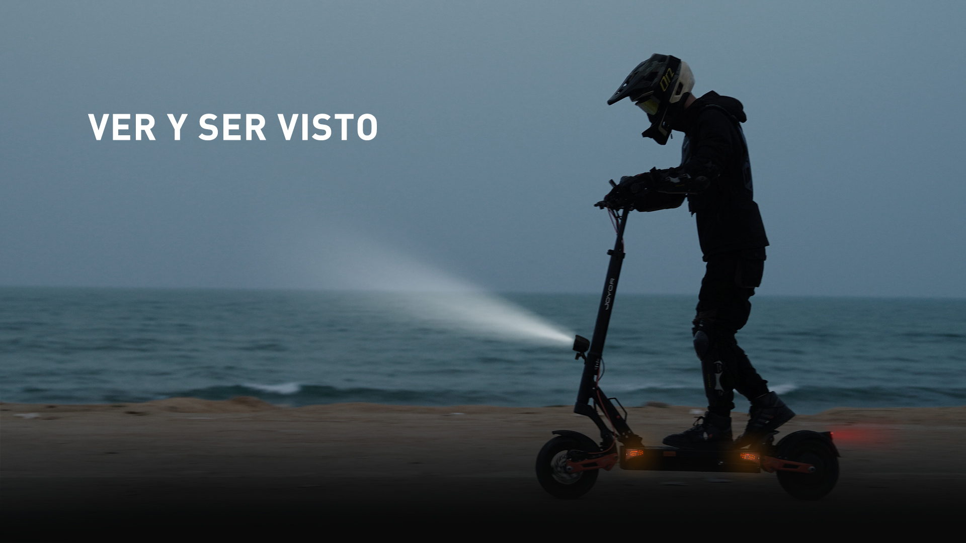 Persona con equipo de protección en patinete eléctrico junto al mar al atardecer.