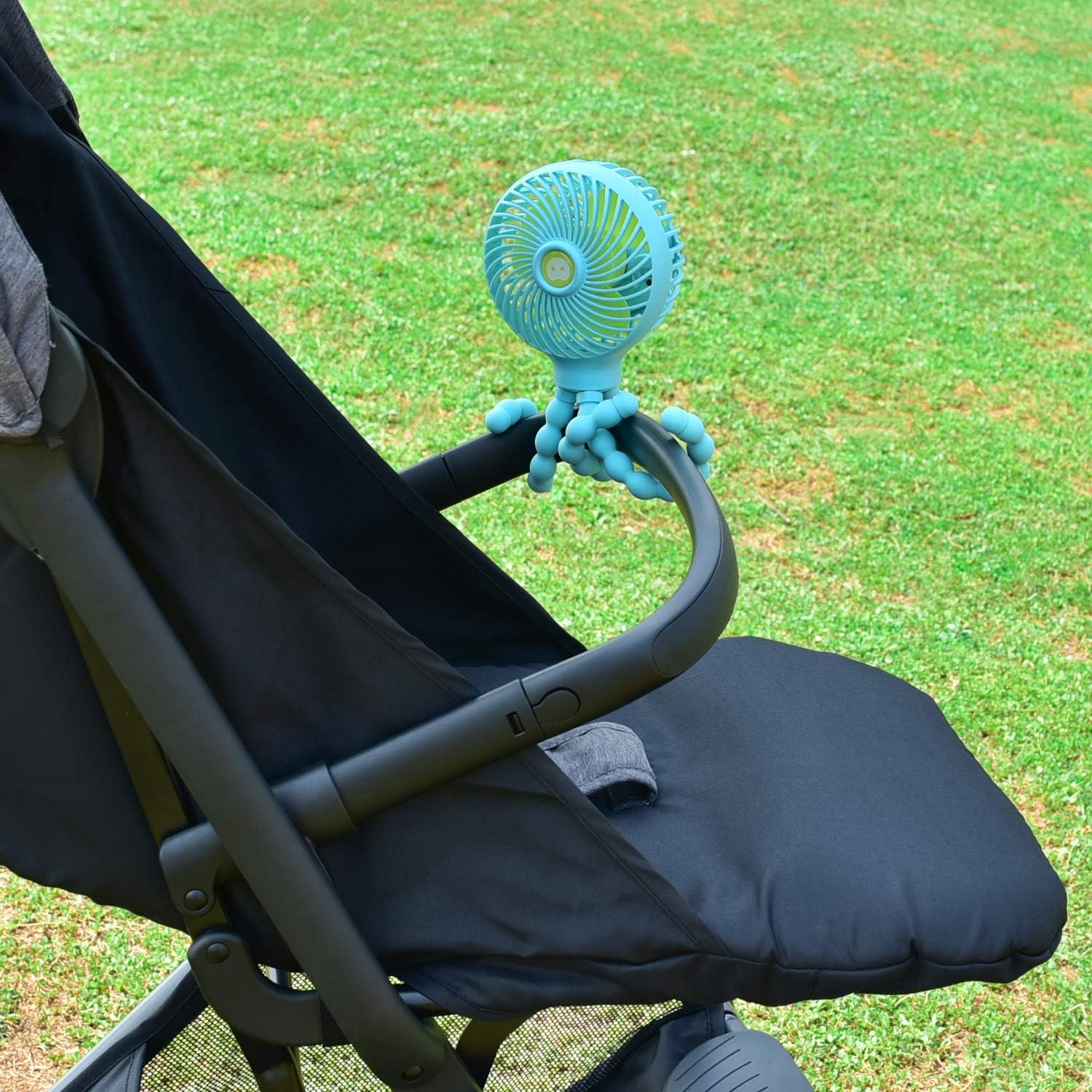 Ein blauer flexibler Ventilator ist am schwarzen Kinderwagengriff befestigt, vor grünem Gras.