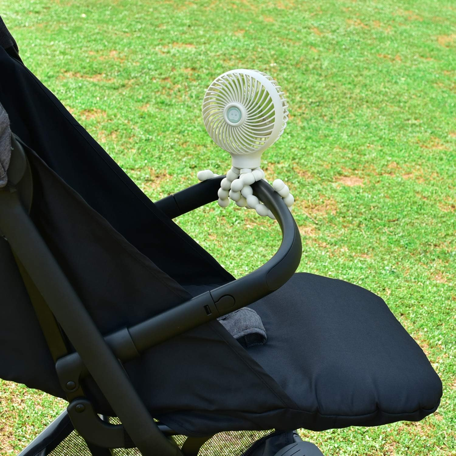 Ein kleiner weißer Ventilator mit flexiblen Beinen ist an einem Kinderwagen befestigt.