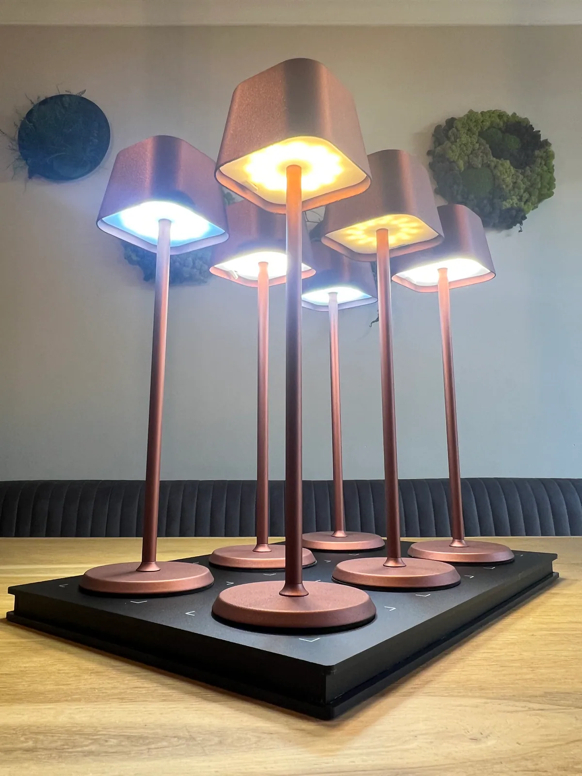 Sechs kupferfarbene Tischlampen mit quadratischen Schirmen stehen auf einer schwarzen Ladestation.