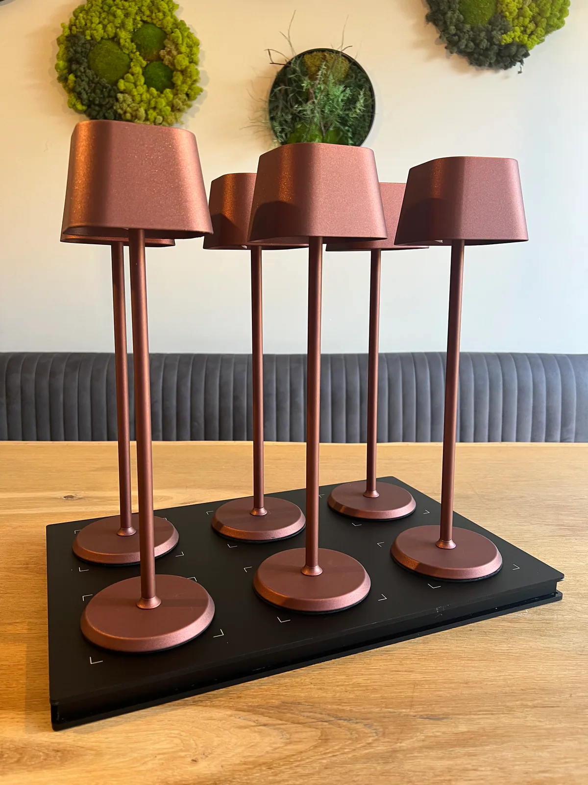 Sechs kupferfarbene Tischlampen stehen auf einem schwarzen Tablett auf einem Holztisch.