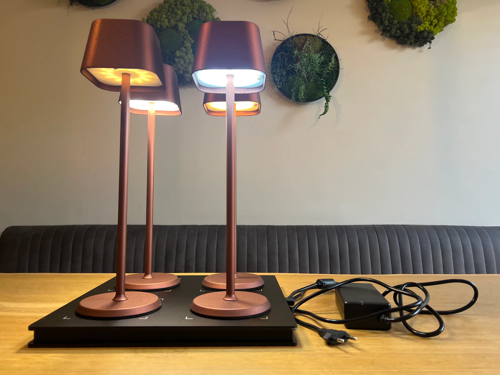 Vier kupferfarbene Tischlampen stehen auf einer schwarzen Ladestation auf einem Holztisch.
