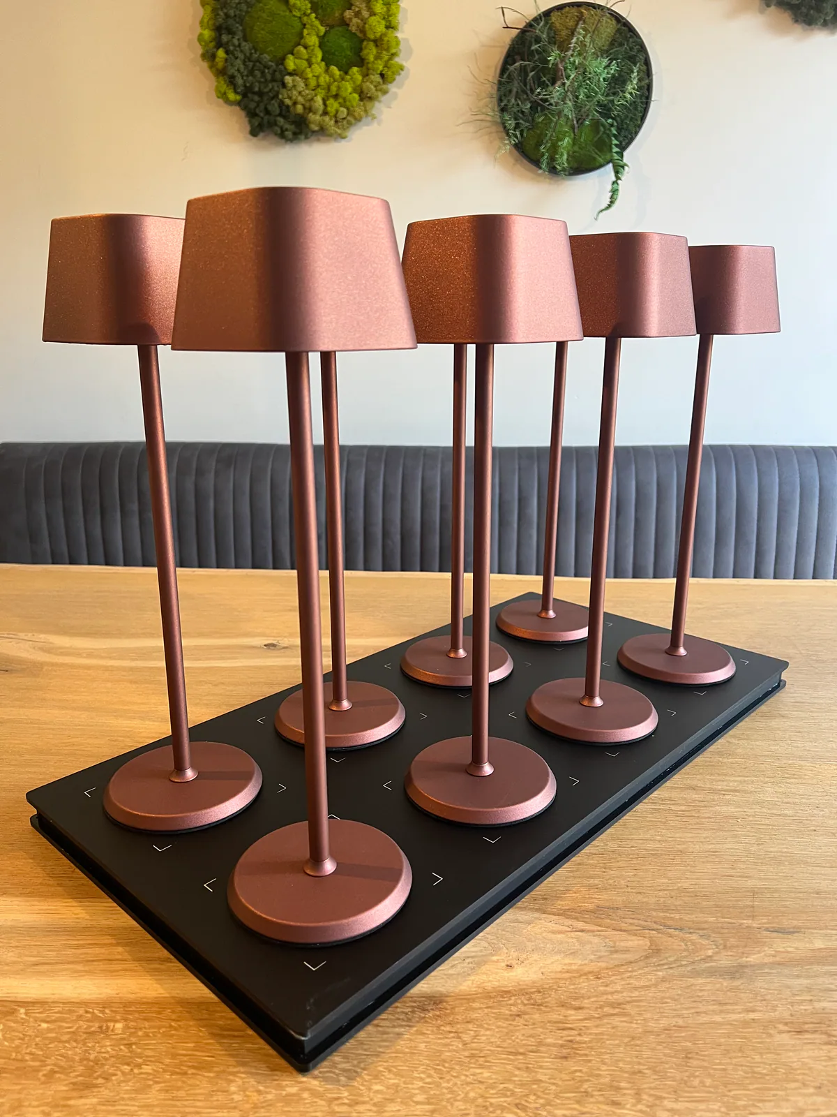 Sechs kupferfarbene Tischlampen stehen auf einer schwarzen Ladestation auf einem Holztisch.