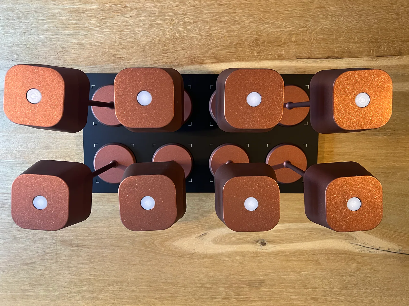 Acht kupferfarbene Würfellichter mit weißen Zentren auf schwarzer Basis, auf Holztisch.