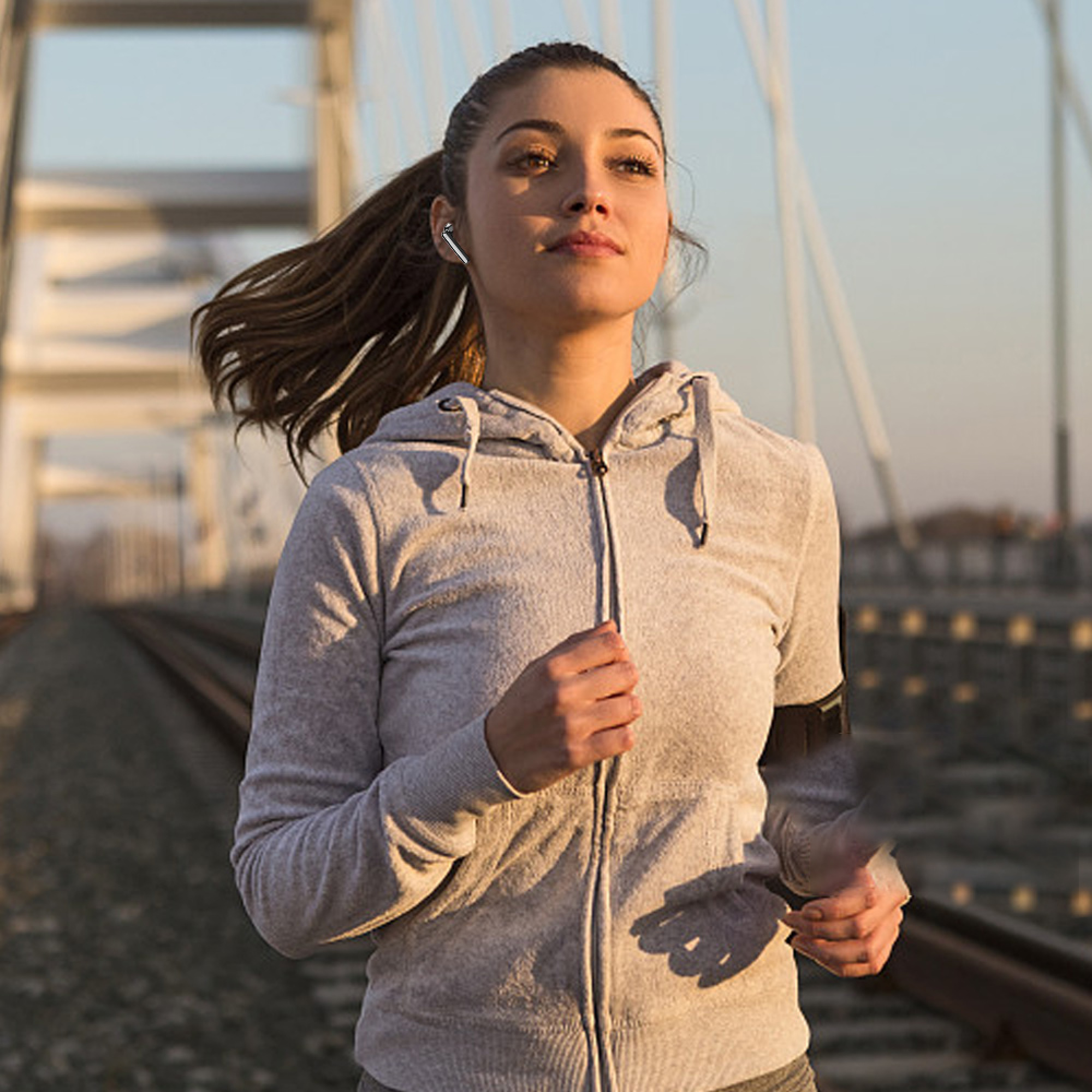 Junge Frau joggt auf einer Brücke über Bahngleise, trägt Kopfhörer und Kapuzenpullover.