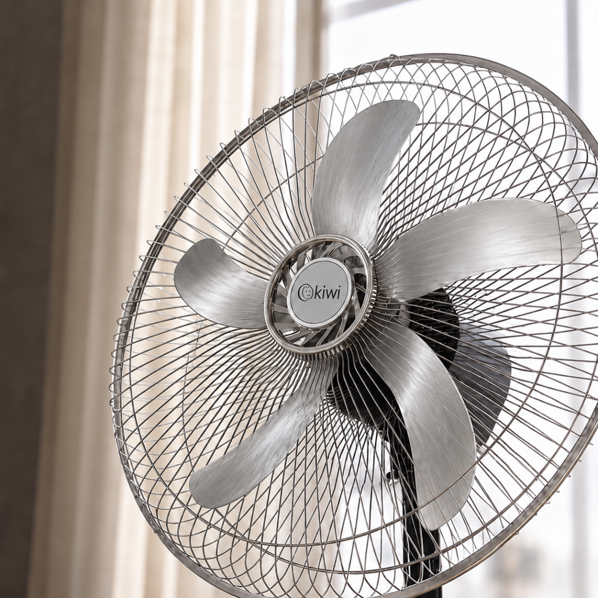 Un ventilatore da terra argentato a cinque pale davanti a una tenda chiara.