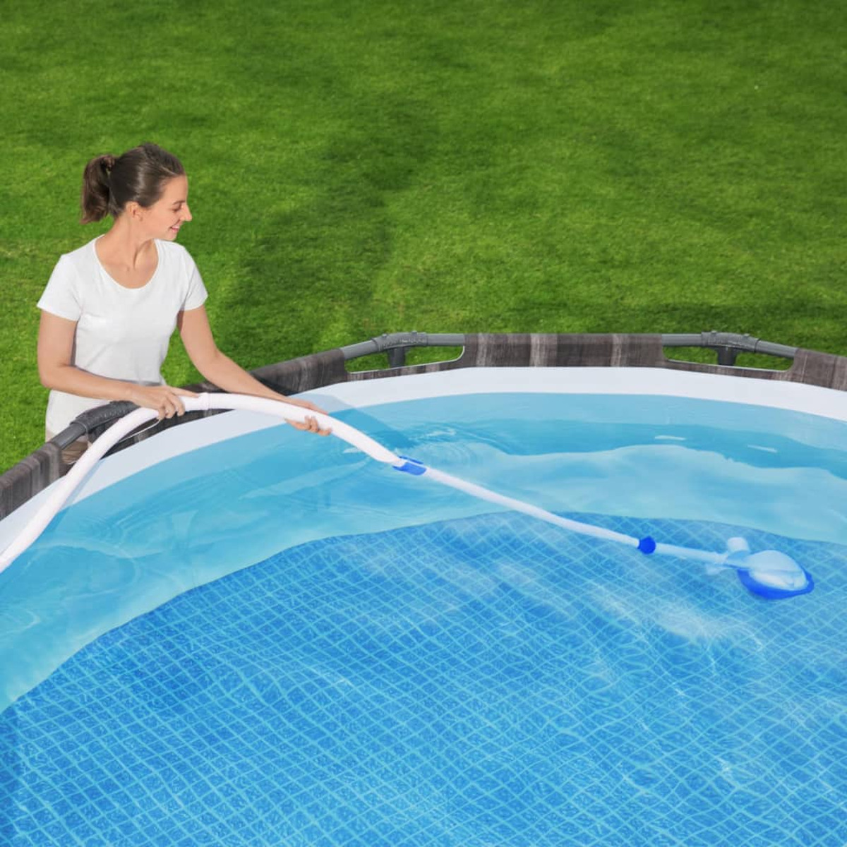 Donna pulisce piscina con aspirapolvere collegato a tubo. Erba verde sullo sfondo.