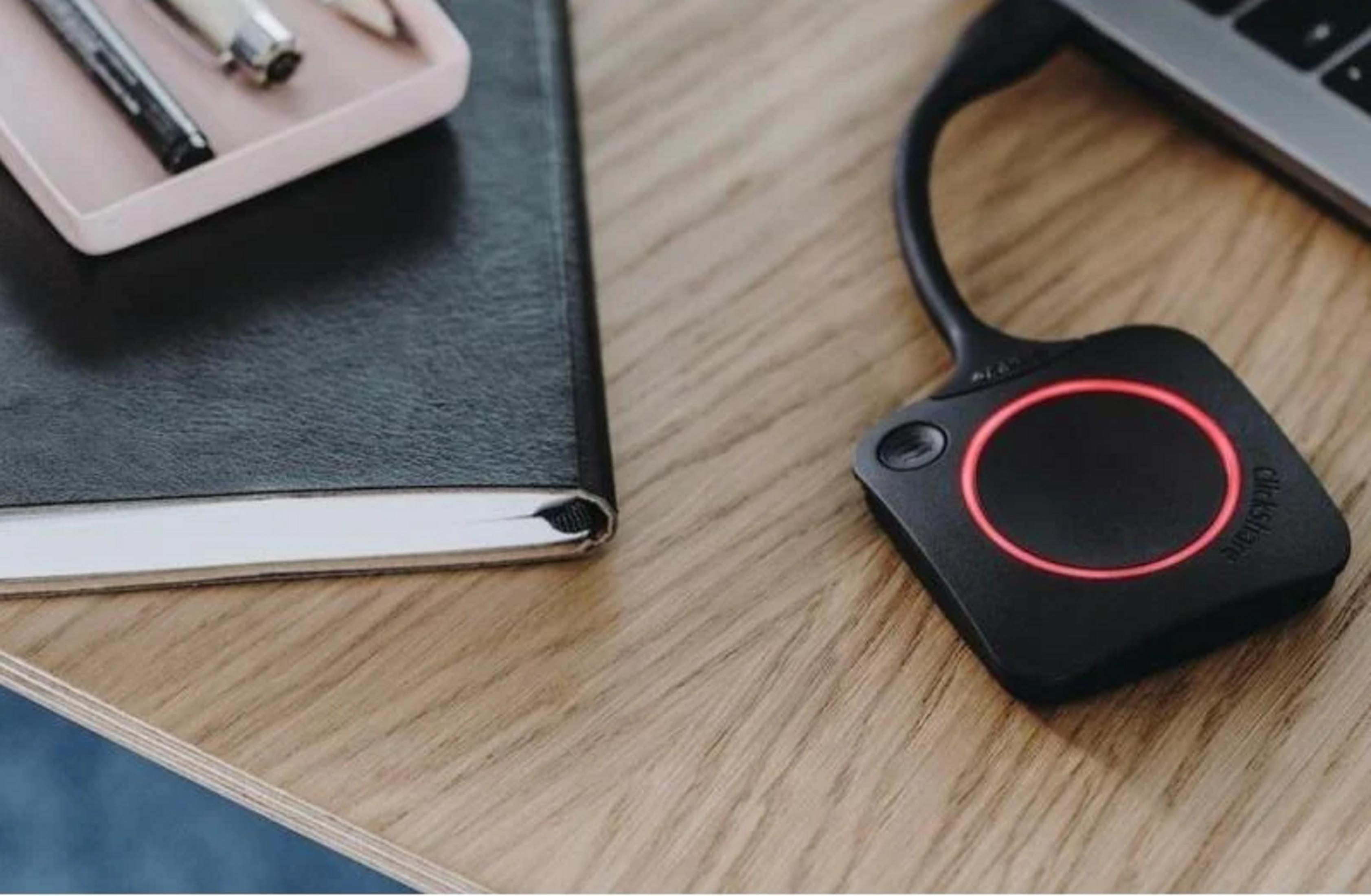 Un trasmettitore ClickShare nero con anello rosso su una scrivania di legno accanto a un taccuino.