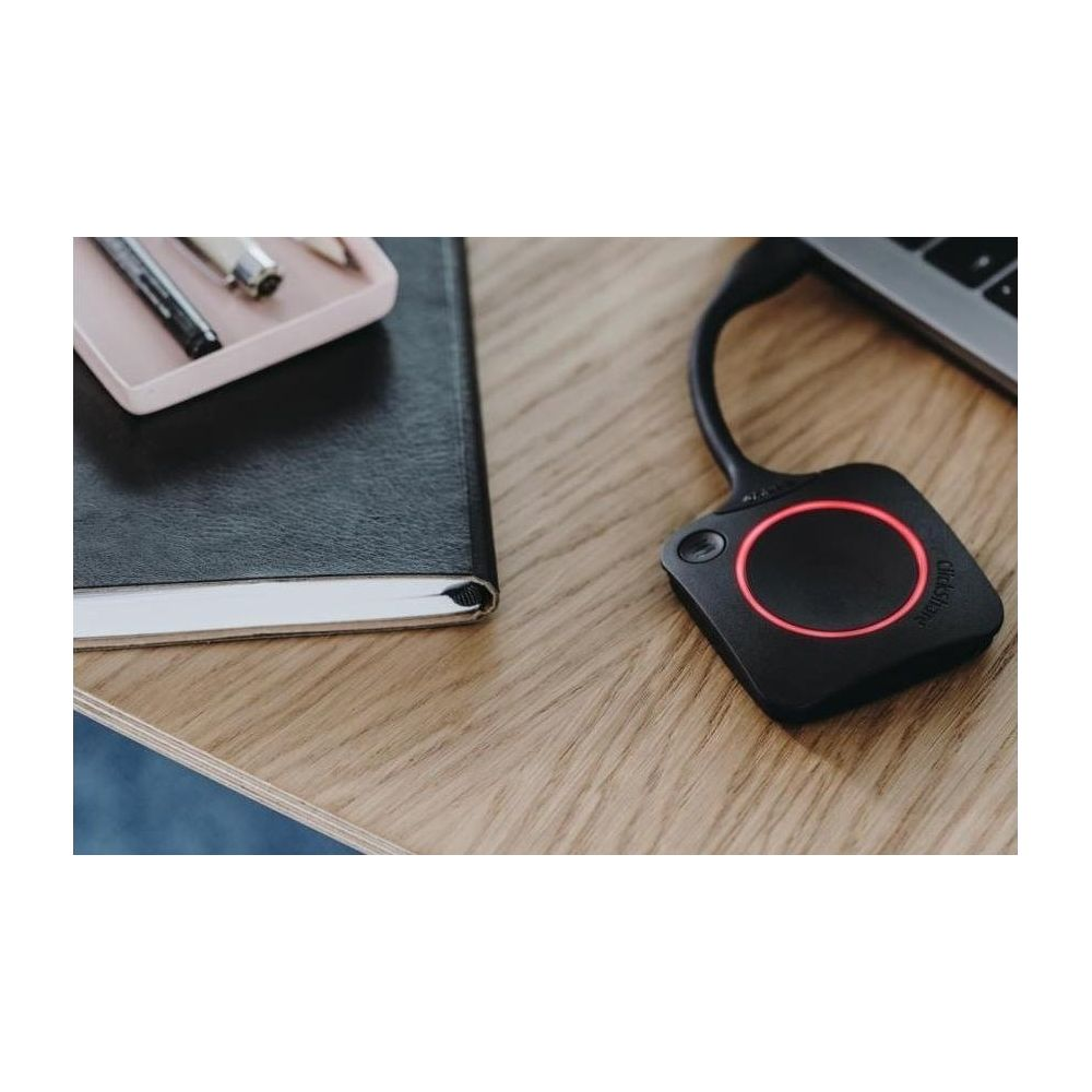 Un trasmettitore ClickShare nero con luce ad anello rossa poggia su una scrivania di legno accanto a un taccuino.