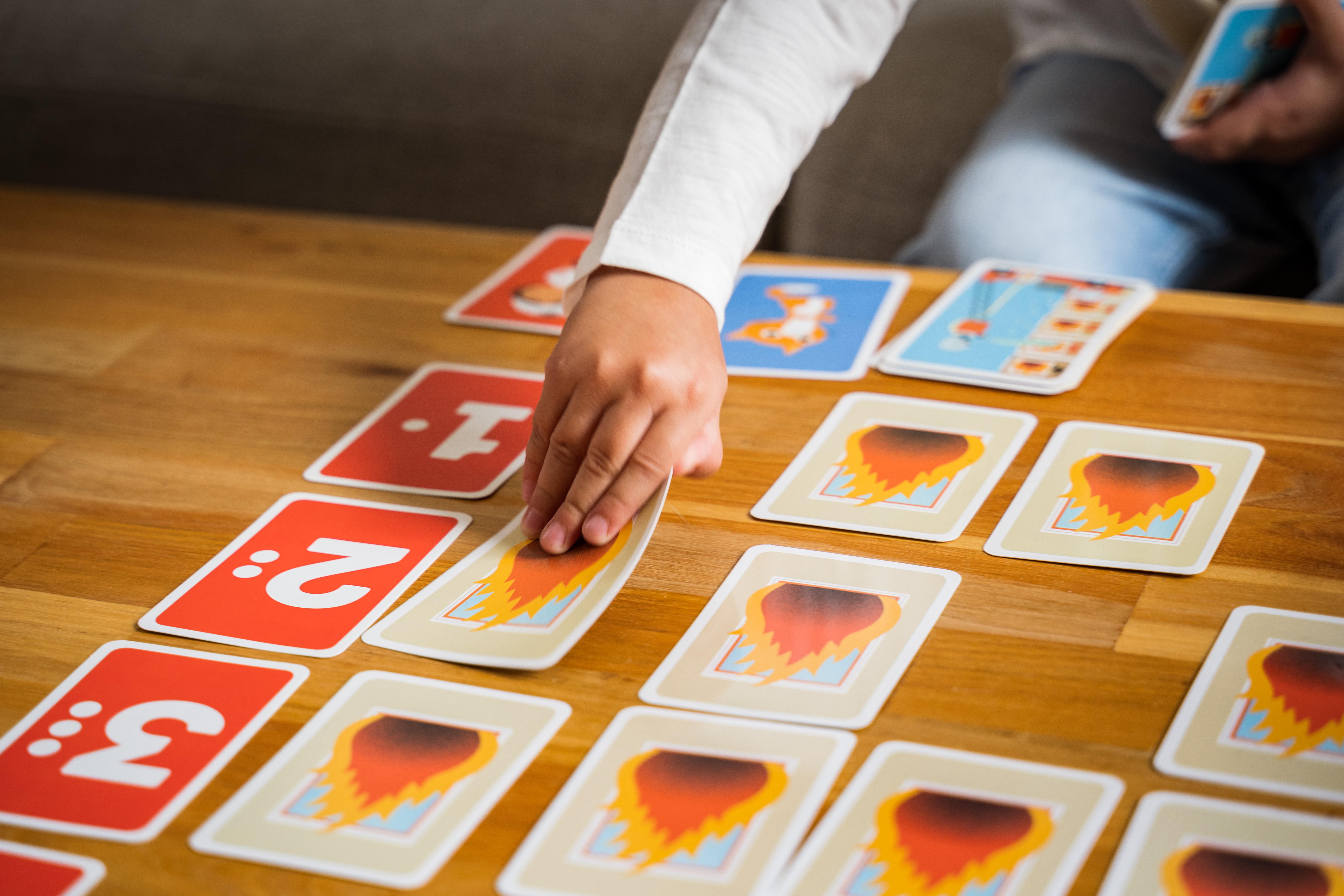 Ein Kind spielt Karten auf einem Holztisch. Einige Karten zeigen Zahlen, andere Flammen.