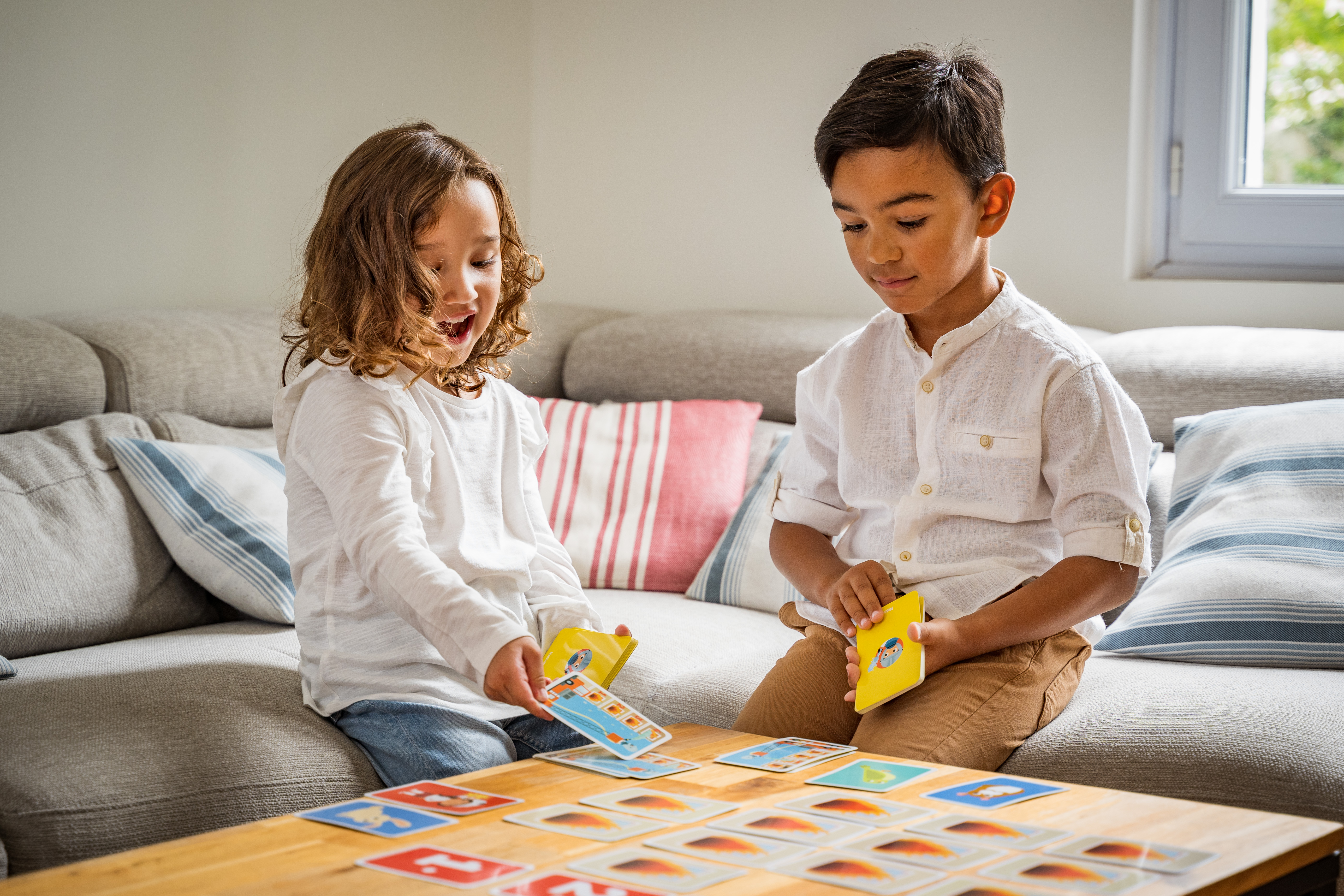 Zwei Kinder spielen Karten auf einem Sofa. Der Tisch ist mit Karten bedeckt.