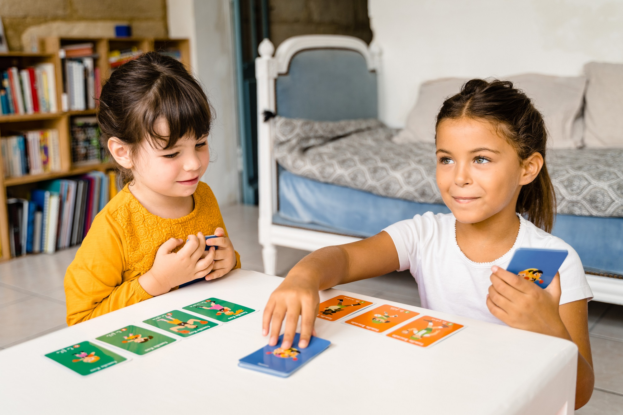 Zwei Mädchen spielen Karten an einem Tisch. Ein Bücherregal und ein Bett sind im Hintergrund.