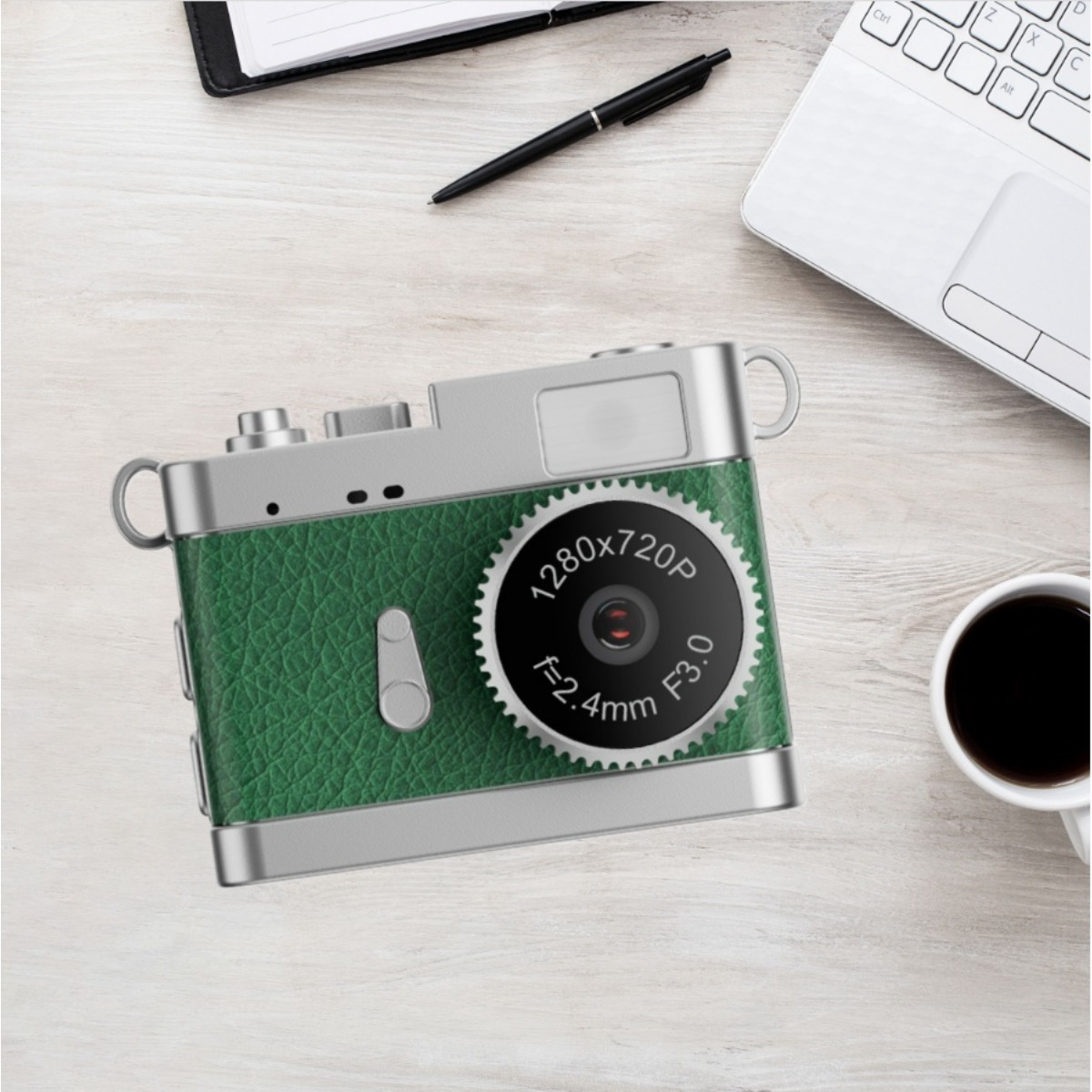 Grüne Kamera mit Silberdetails, auf einem Holztisch mit Laptop, Stift und Kaffee.
