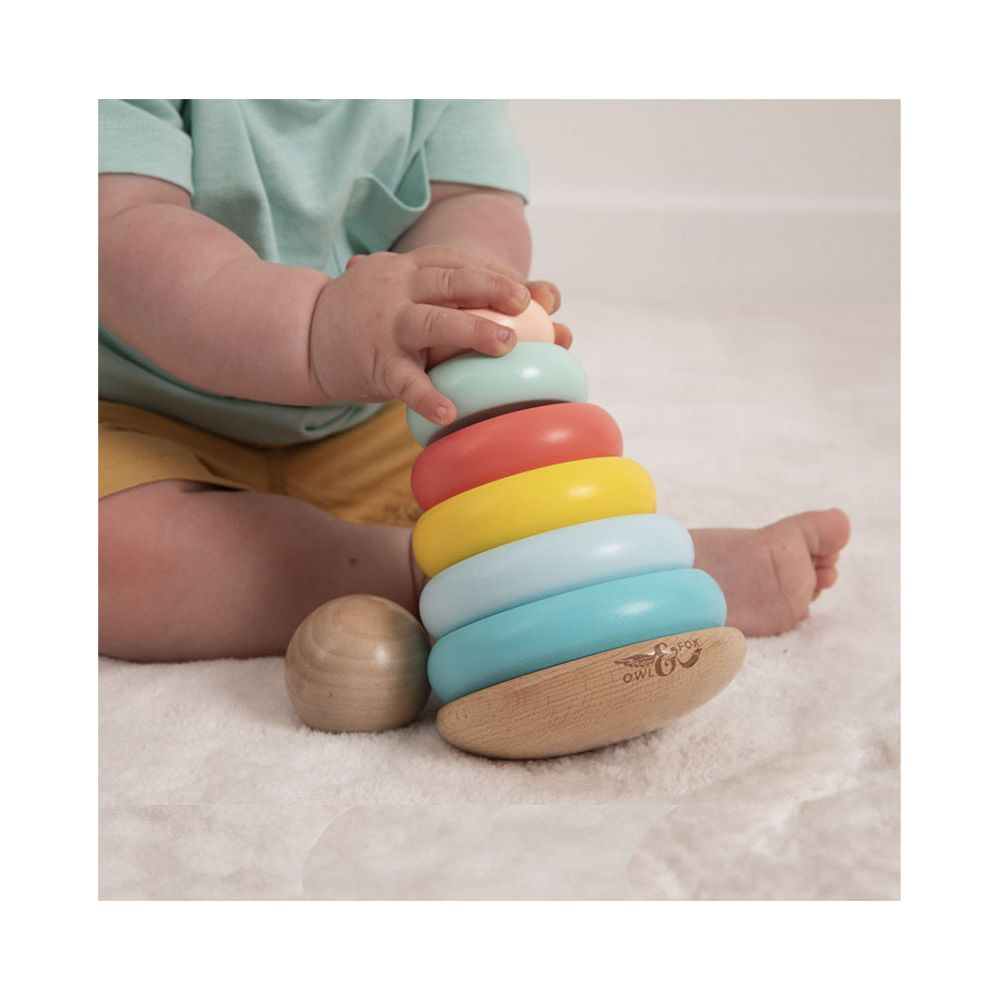 Ein Baby spielt mit einem Holzringturm in Pastellfarben auf einem flauschigen Teppich.