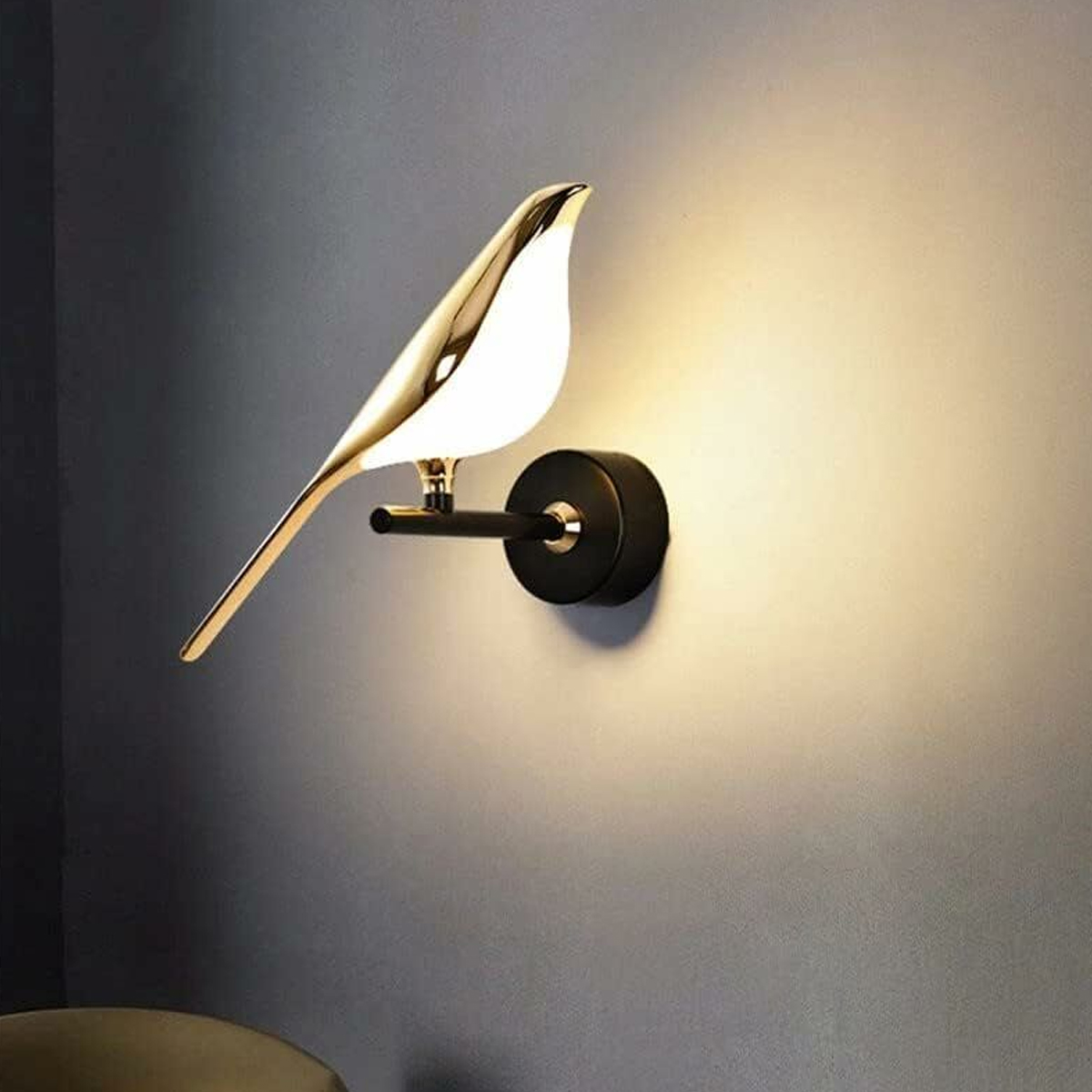 Eine goldene vogelformige Wandleuchte mit schwarzem Sockel ist an einer grauen Wand montiert.