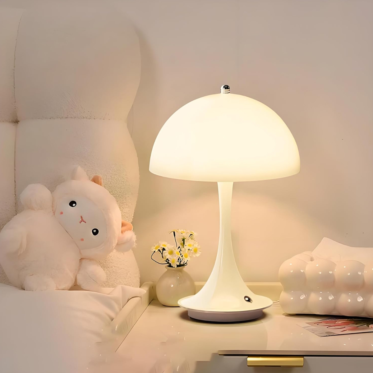 Ein weißer Nachttisch mit Lampe, Lamm-Plüschtier und Blumen in Vase.