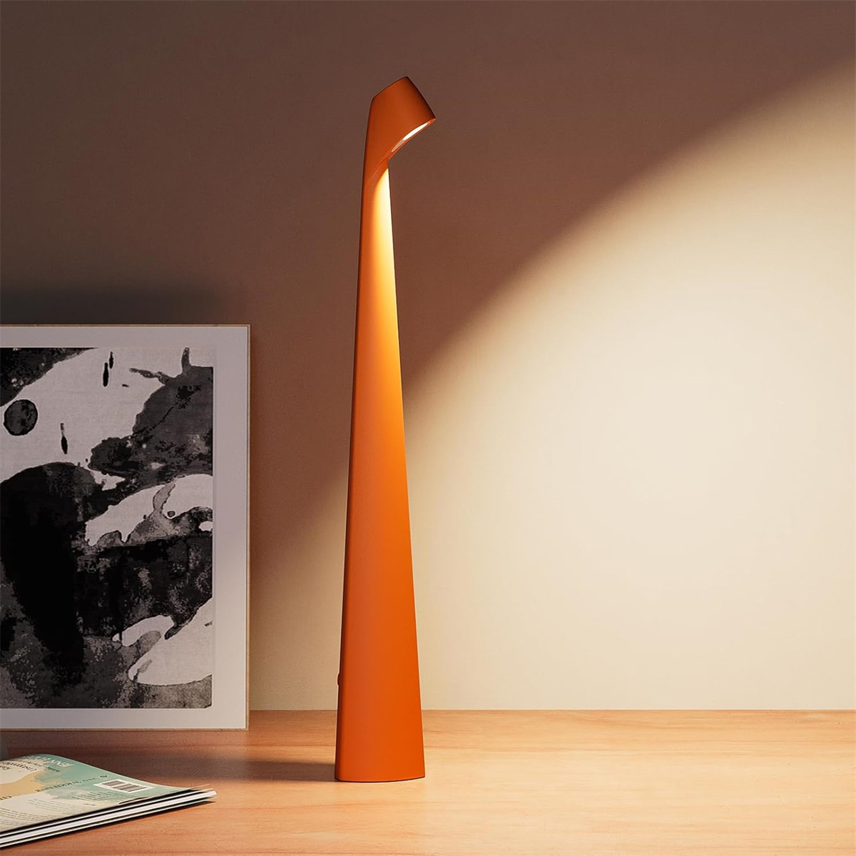 Eine schmale, orange Stehlampe steht auf einem Holztisch neben einem gerahmten Bild.