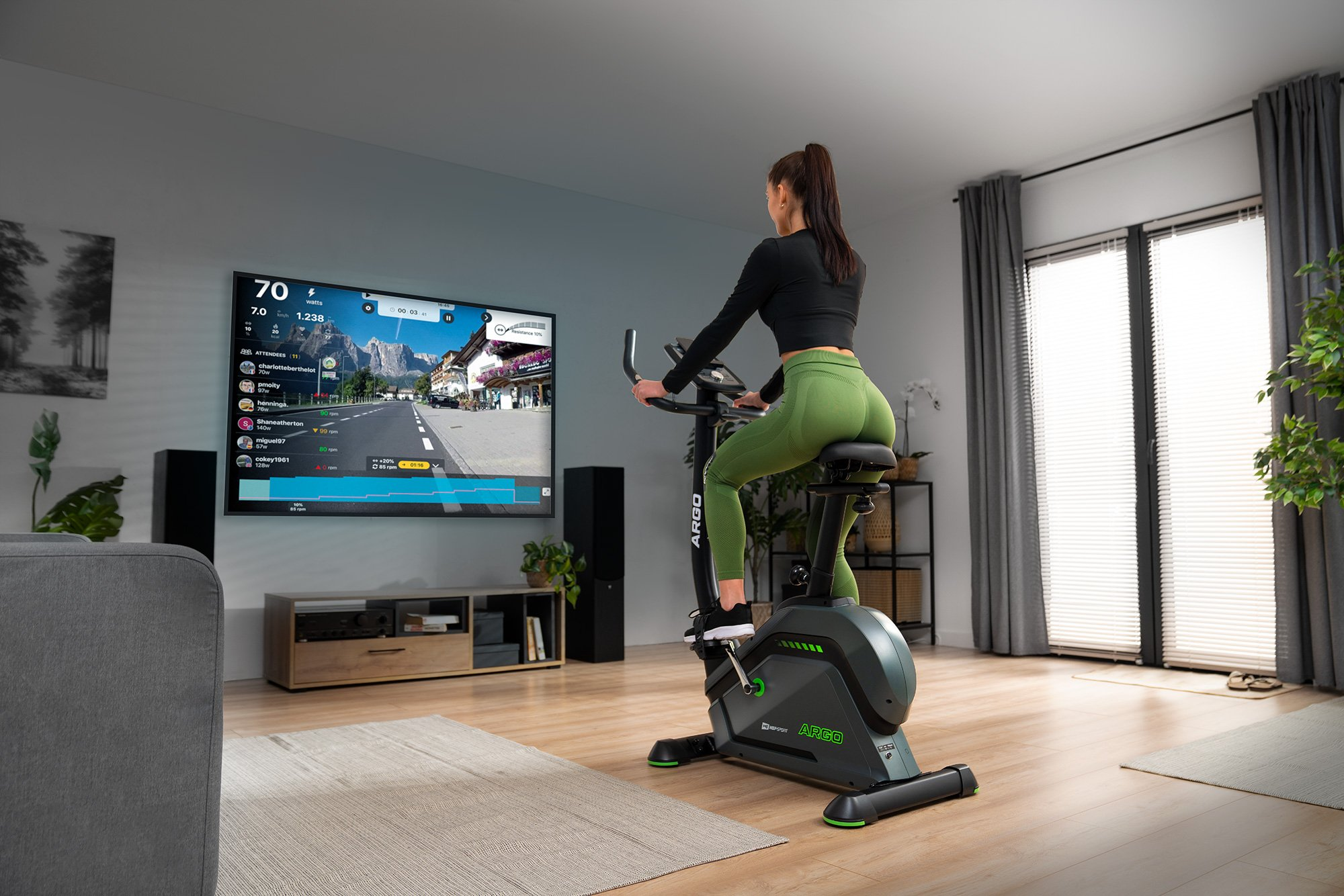 Eine Frau trainiert auf einem Heimtrainer vor einem großen Bildschirm, der eine virtuelle Fahrt zeigt.