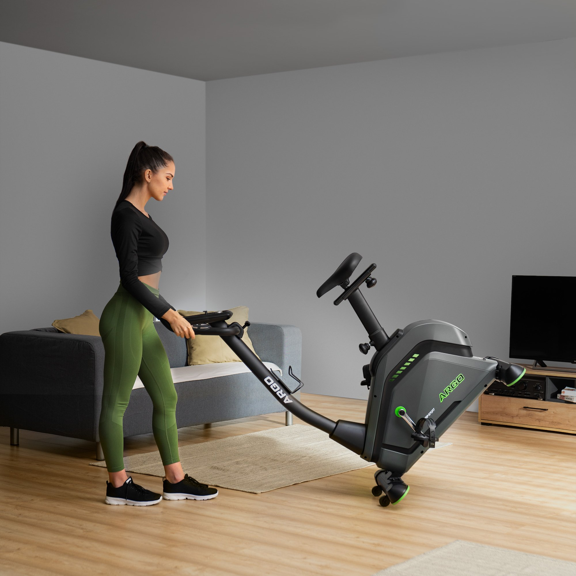 Eine Frau in Trainingskleidung steht neben einem Heimtrainer in einem Wohnzimmer.