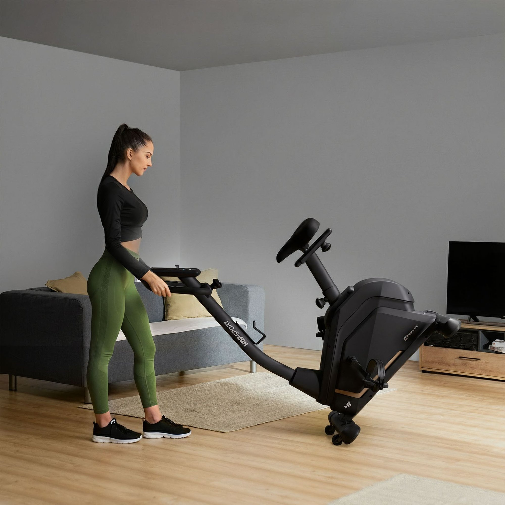 Frau in Sportkleidung steht neben einem schwarzen Heimtrainer in einem Wohnzimmer.