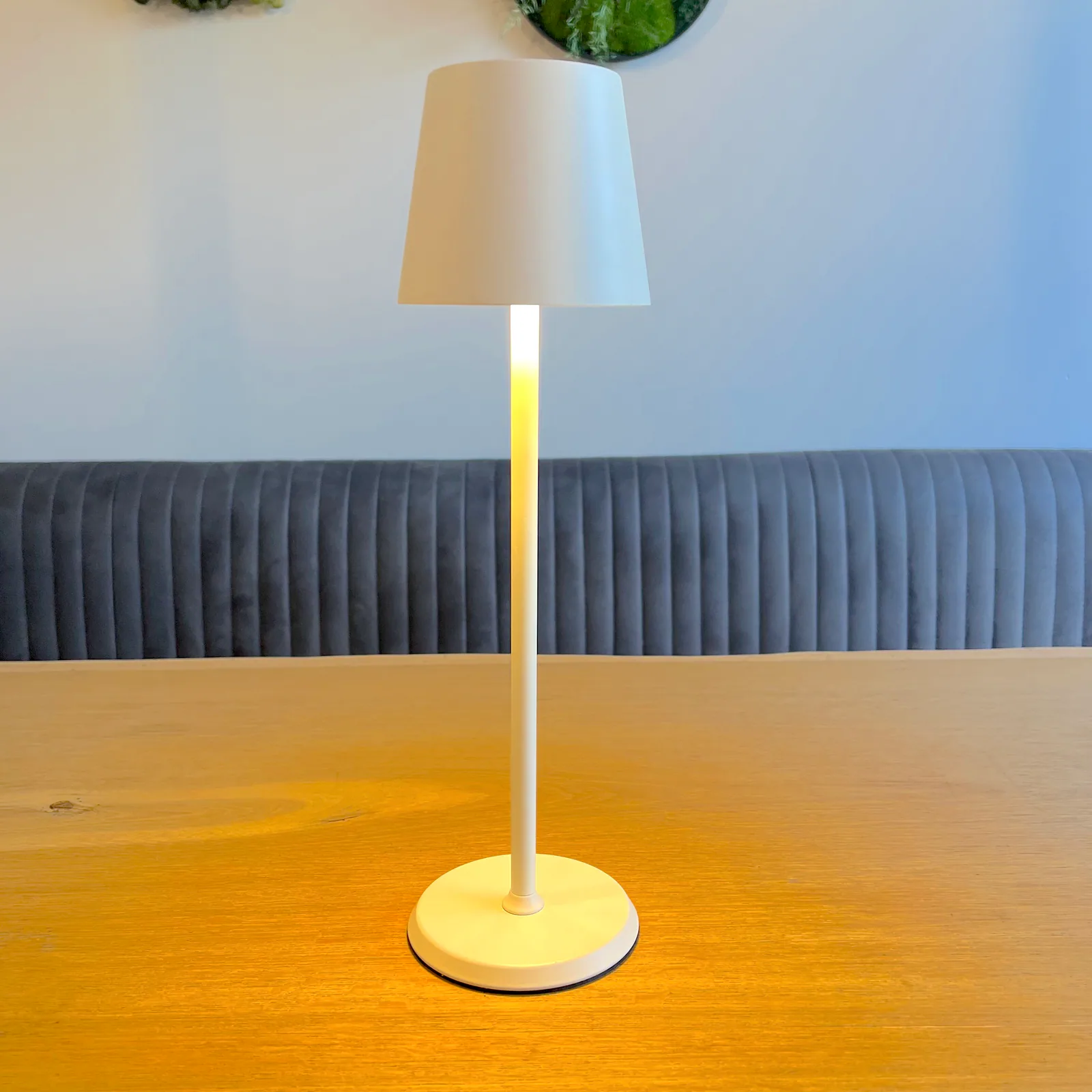 Eine weiße Lampe steht auf einem Holztisch vor einer grauen Sitzbank.