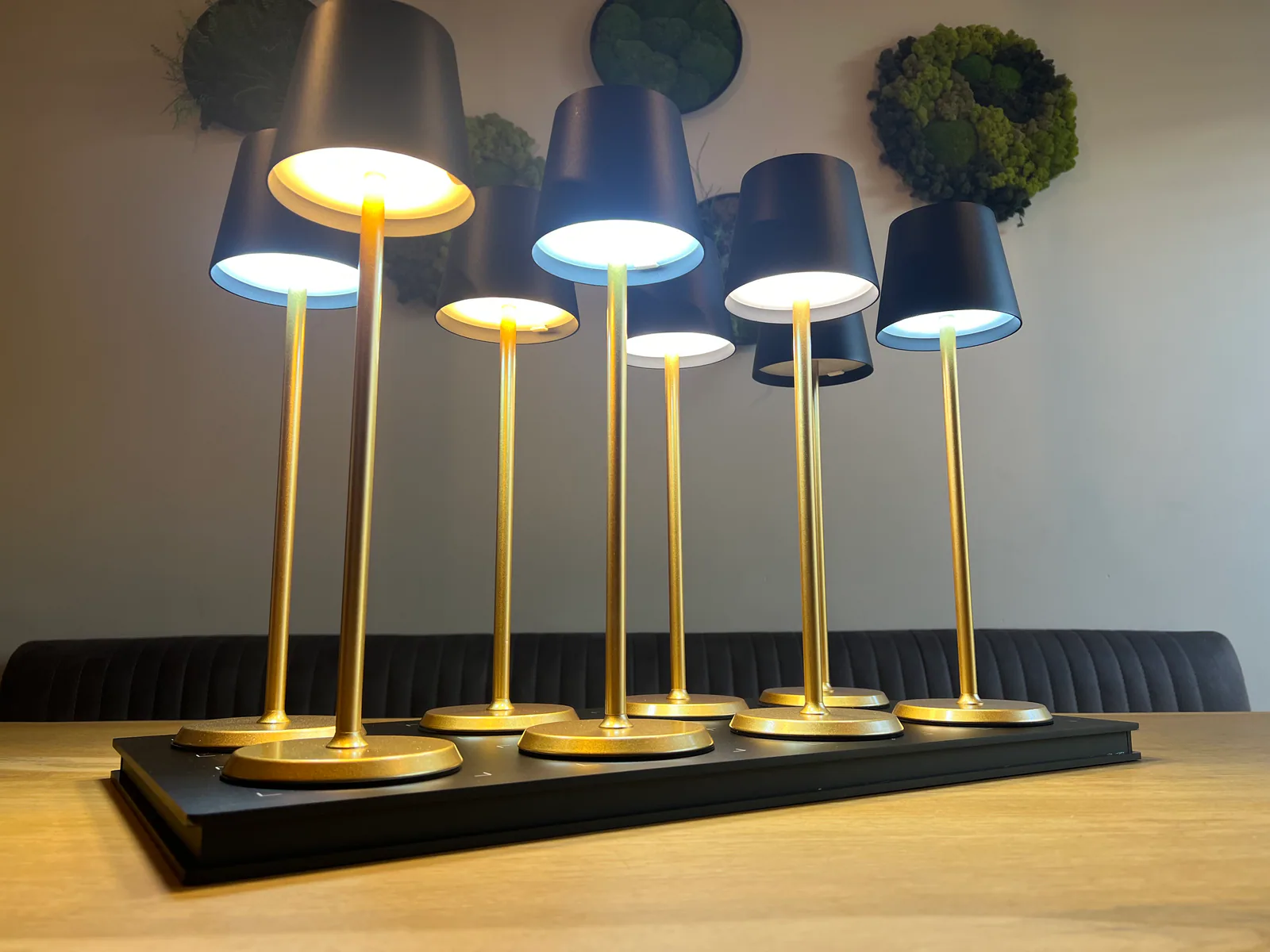Sieben goldene Tischlampen mit schwarzen Schirmen auf einem schwarzen Sockel.