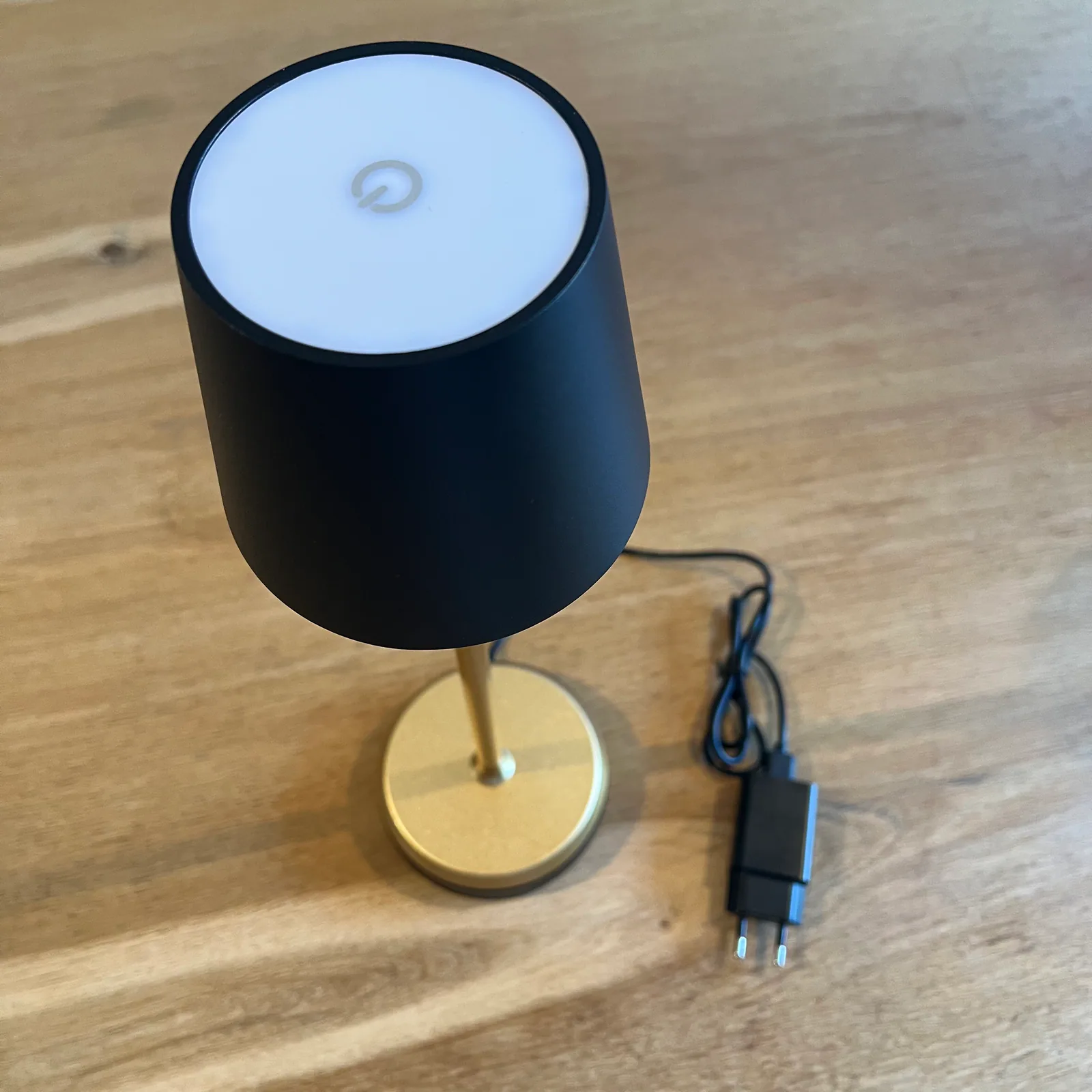 Schwarze Lampe mit goldenem Sockel und Netzteil auf Holztisch.