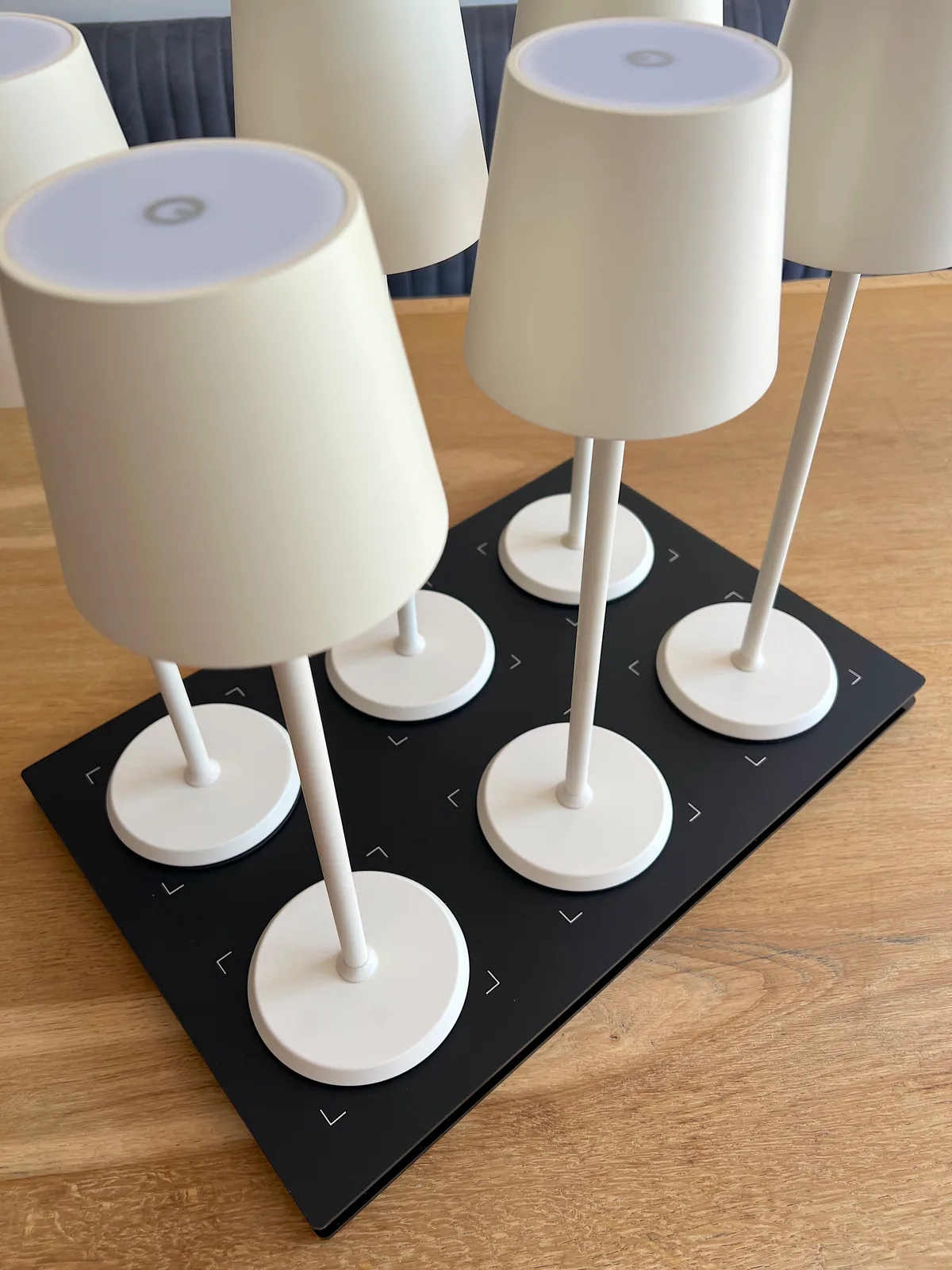 Sechs weiße Lampen stehen auf einer schwarzen Ladestation auf einem Holztisch.