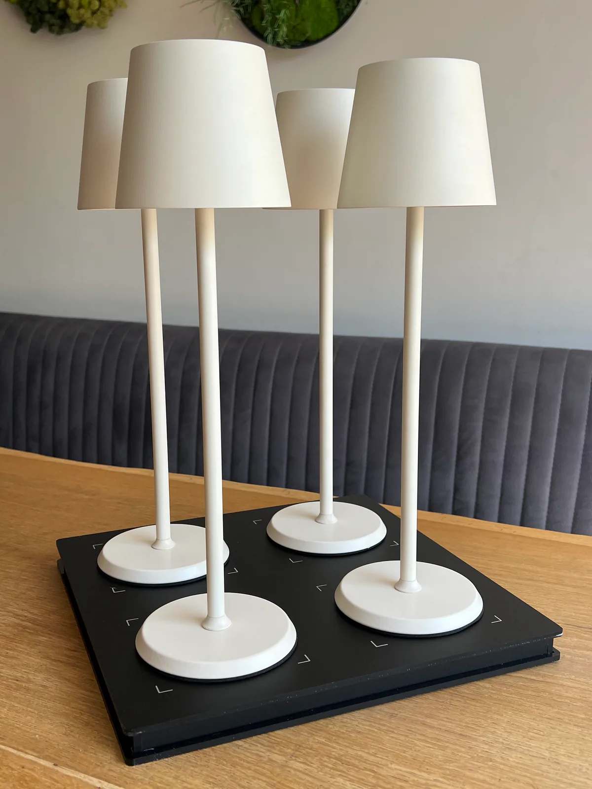 Vier weiße Tischlampen stehen auf einer schwarzen Ladestation auf einem Holztisch.