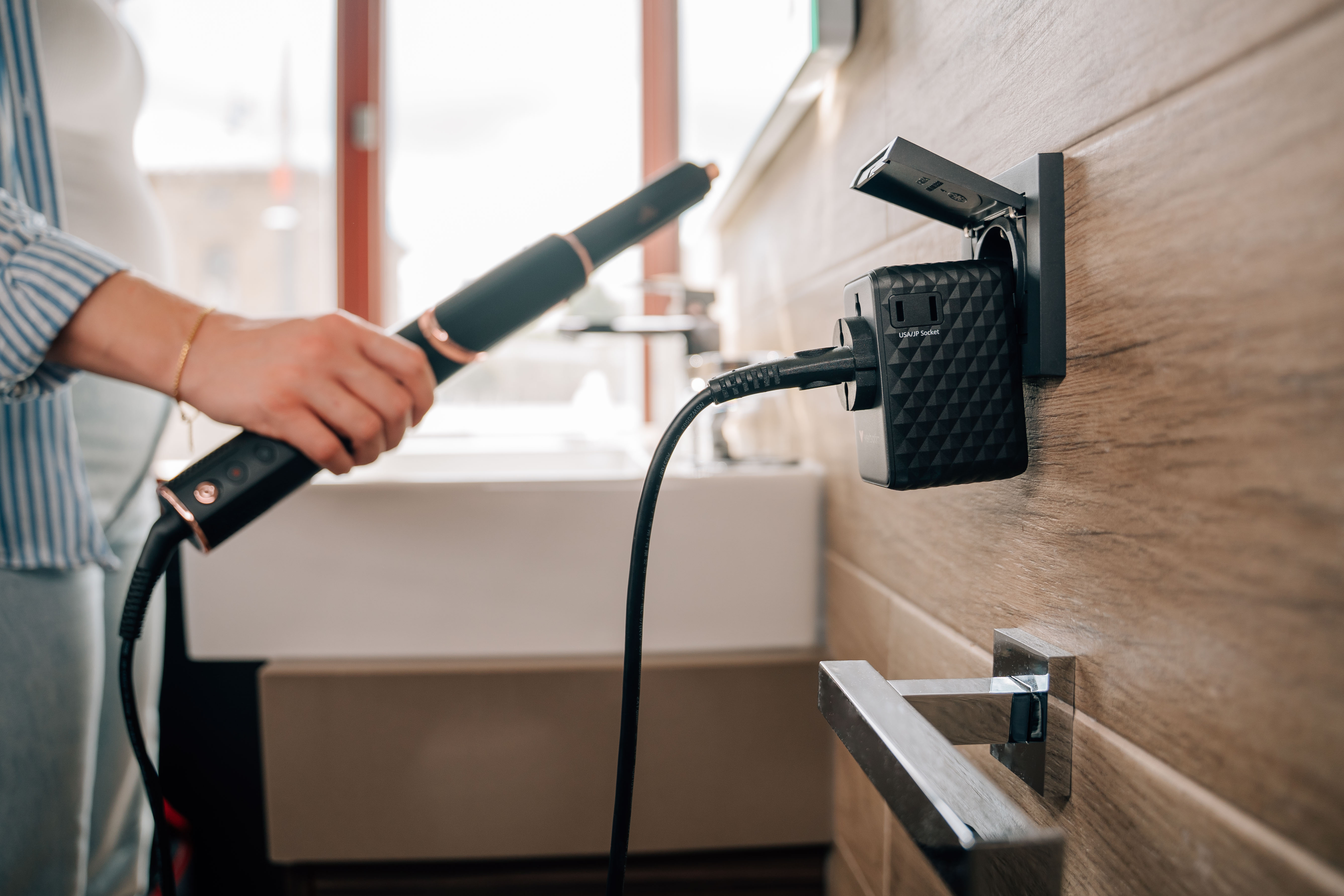 Une personne branche un fer à friser noir dans un adaptateur de voyage universel sur un mur en bois.