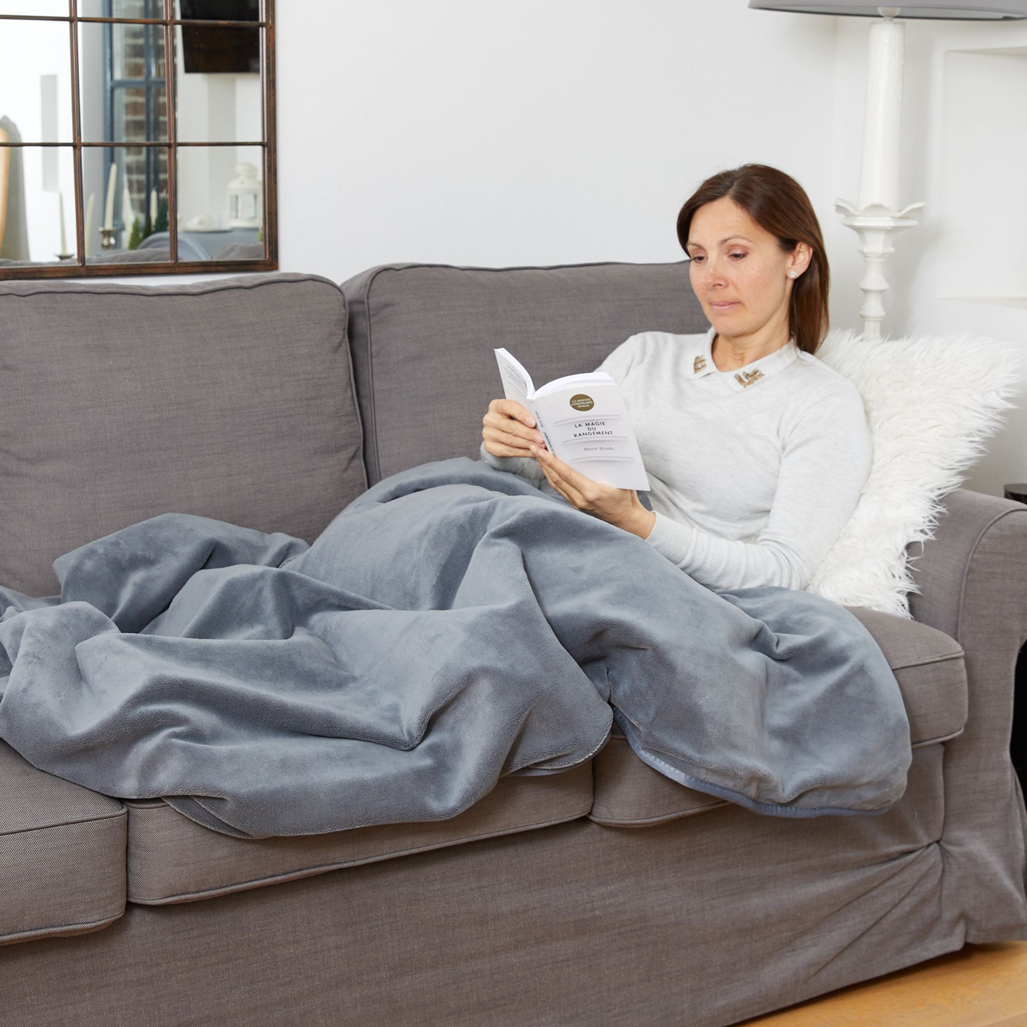 Frau liest Buch auf grauem Sofa unter einer Decke, mit Spiegel und Lampe im Hintergrund.