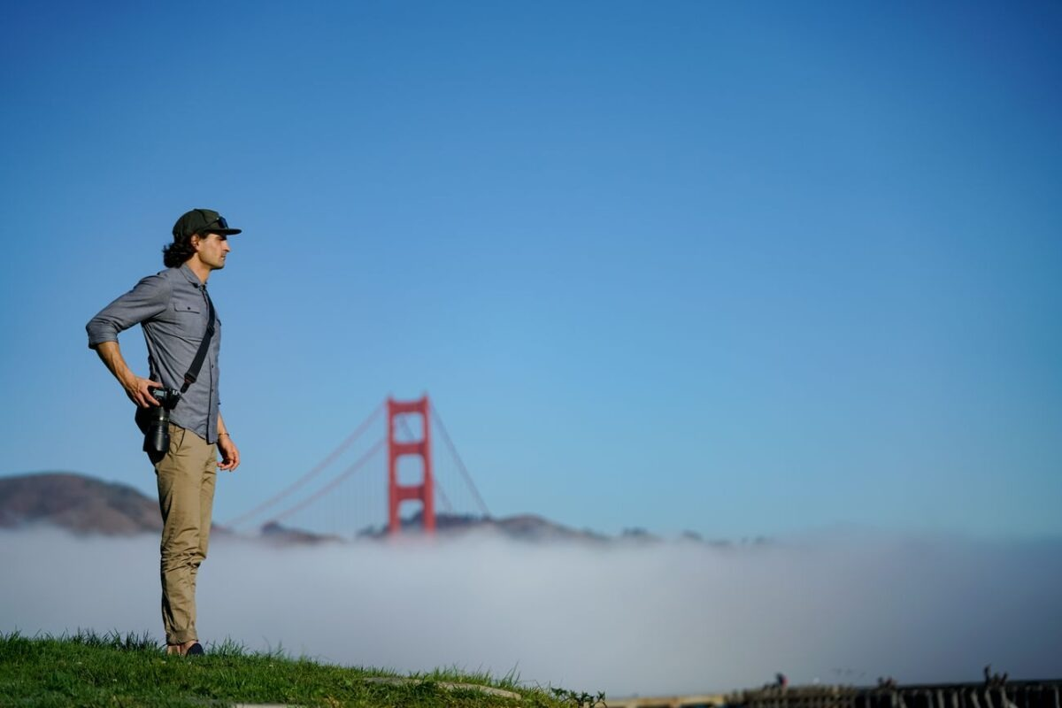 Uomo con macchina fotografica davanti al Golden Gate Bridge nebbioso sotto cielo blu.