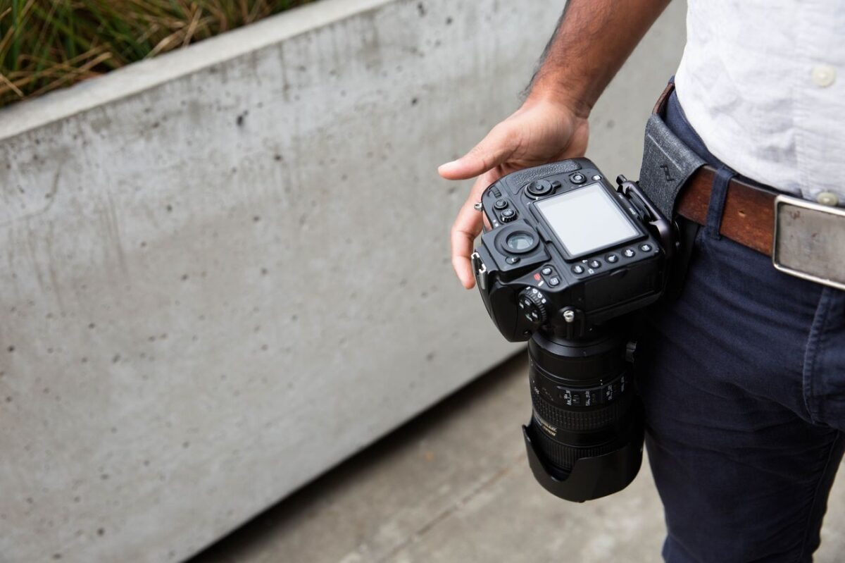 Uomo in pantaloni scuri e camicia bianca tiene una fotocamera DSLR nera con obiettivo grande agganciata alla cintura.