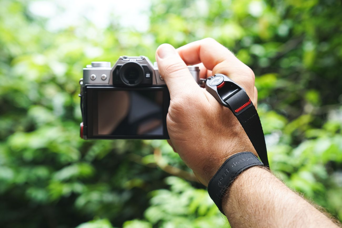 Una mano tiene una fotocamera argentata con cinturino nero, su fogliame verde sfocato.