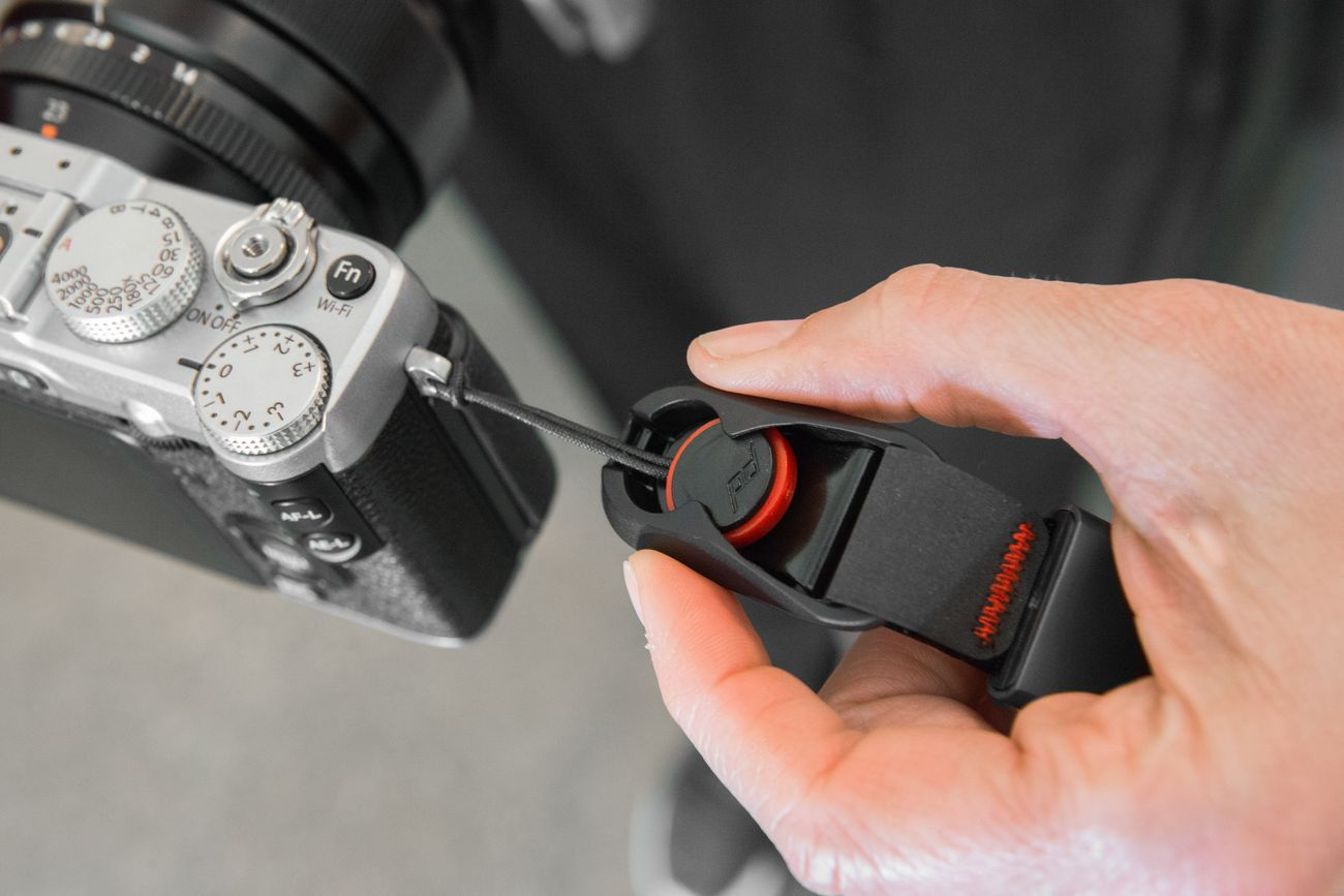Una mano attacca un cinturino per fotocamera nero con dettaglio rosso a una fotocamera argentata.