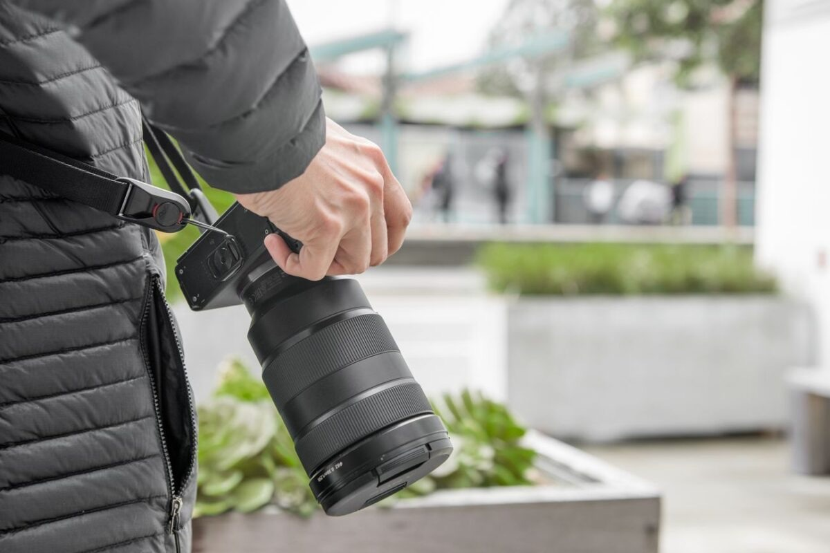 Una mano tiene una fotocamera con un obiettivo grande, con una scena urbana sfocata sullo sfondo.