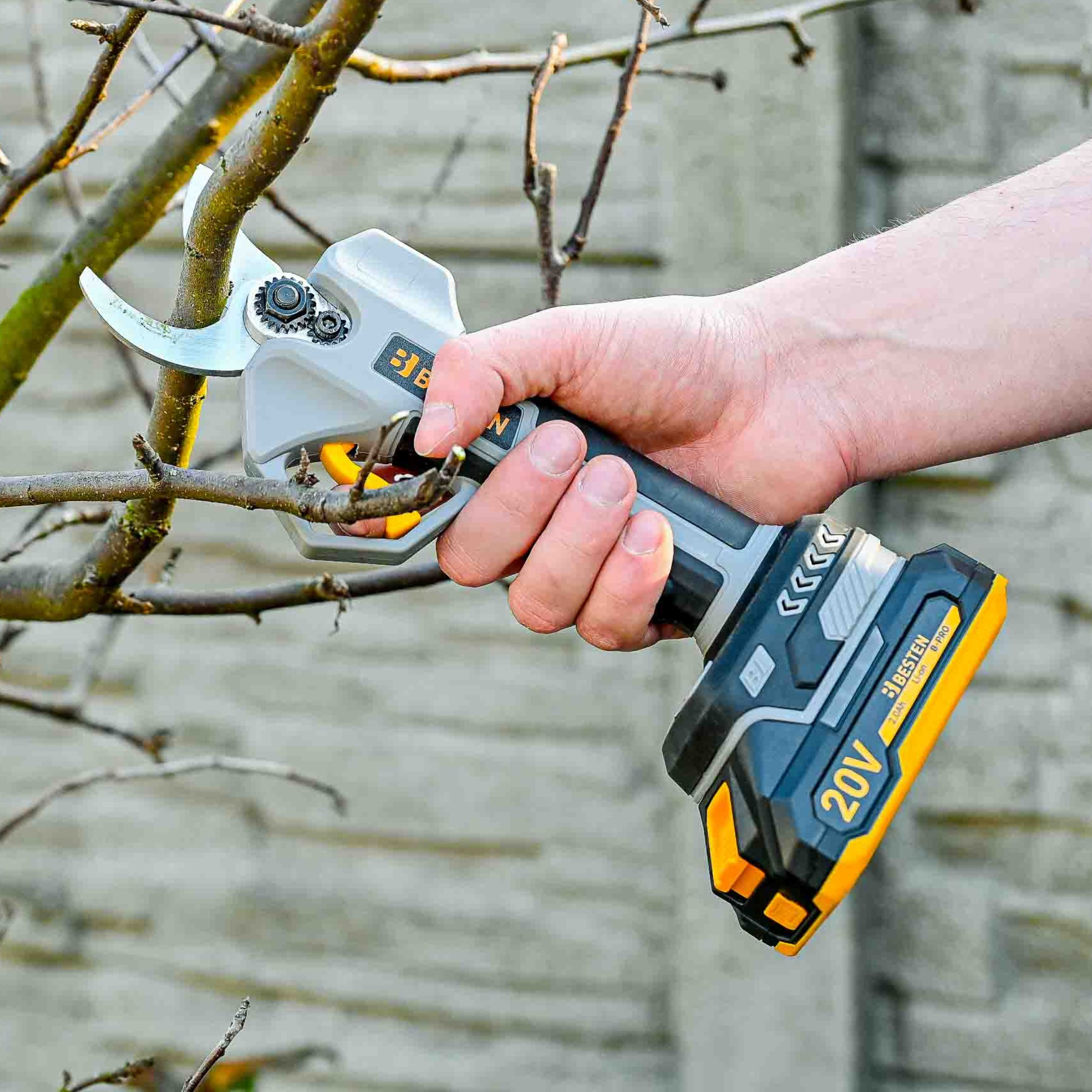 Dłoń trzyma elektryczne nożyce do przycinania na gałęzi drzewa. Szare tło.