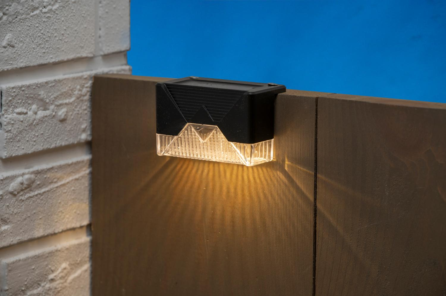 Black solar light on a wooden fence, illuminating the fence with yellow light.