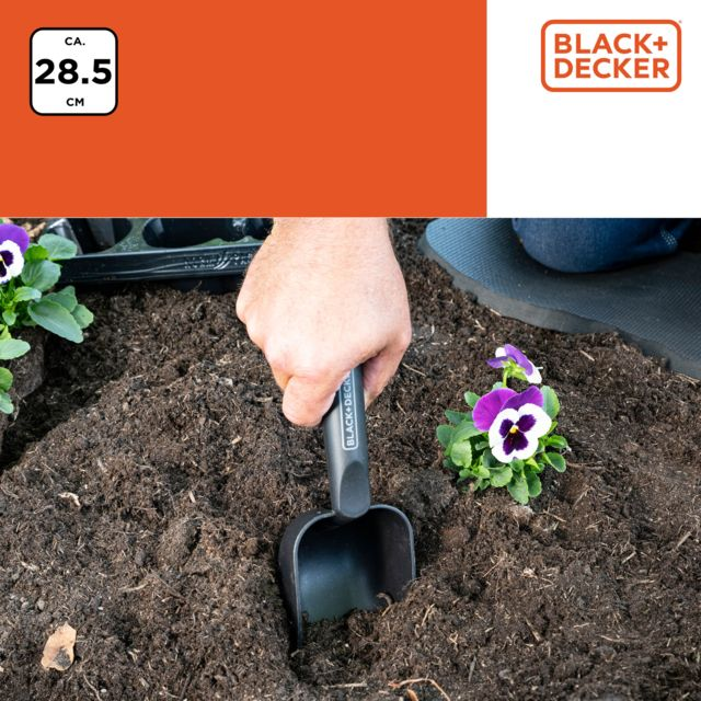 A hand uses a small black trowel to loosen soil around a purple and white flower.