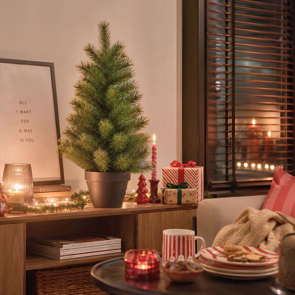 A small Christmas tree sits on a wooden shelf with gifts and candles. Cozy room.
