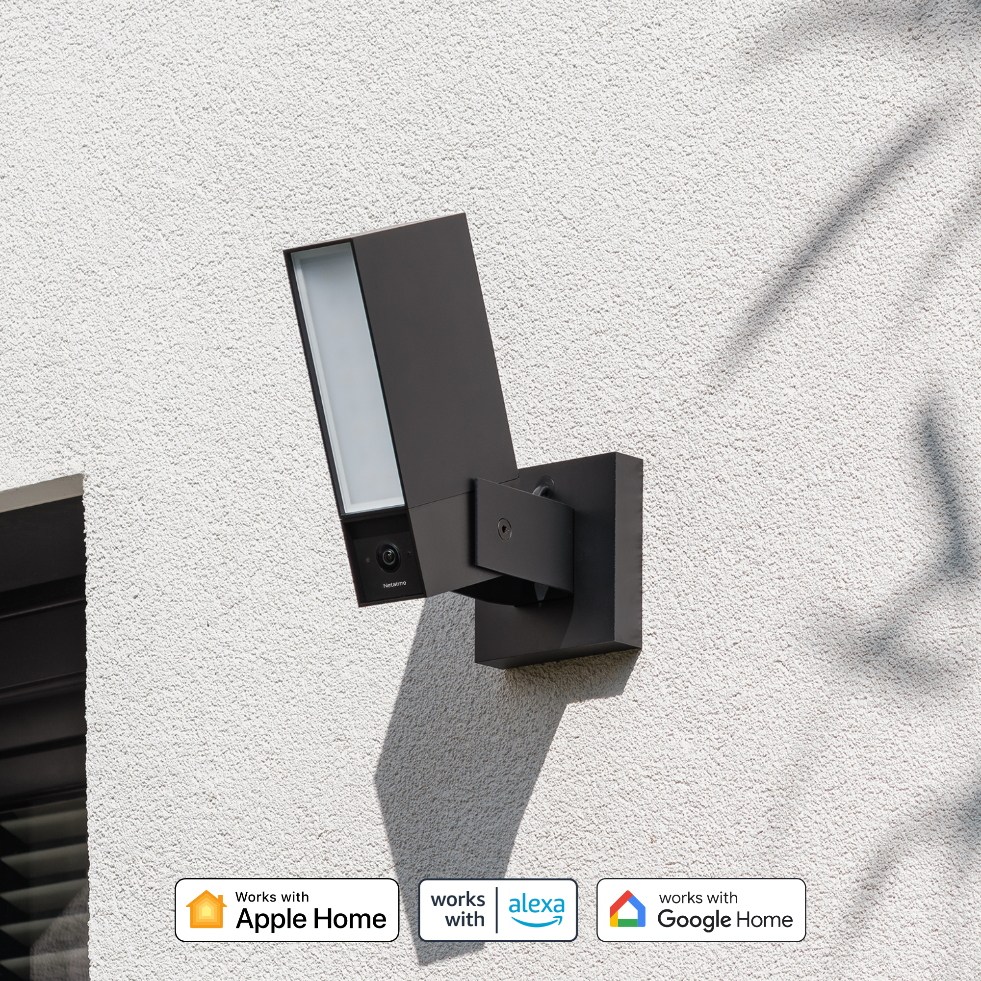 A black outdoor smart camera with a light is mounted on a textured white wall.