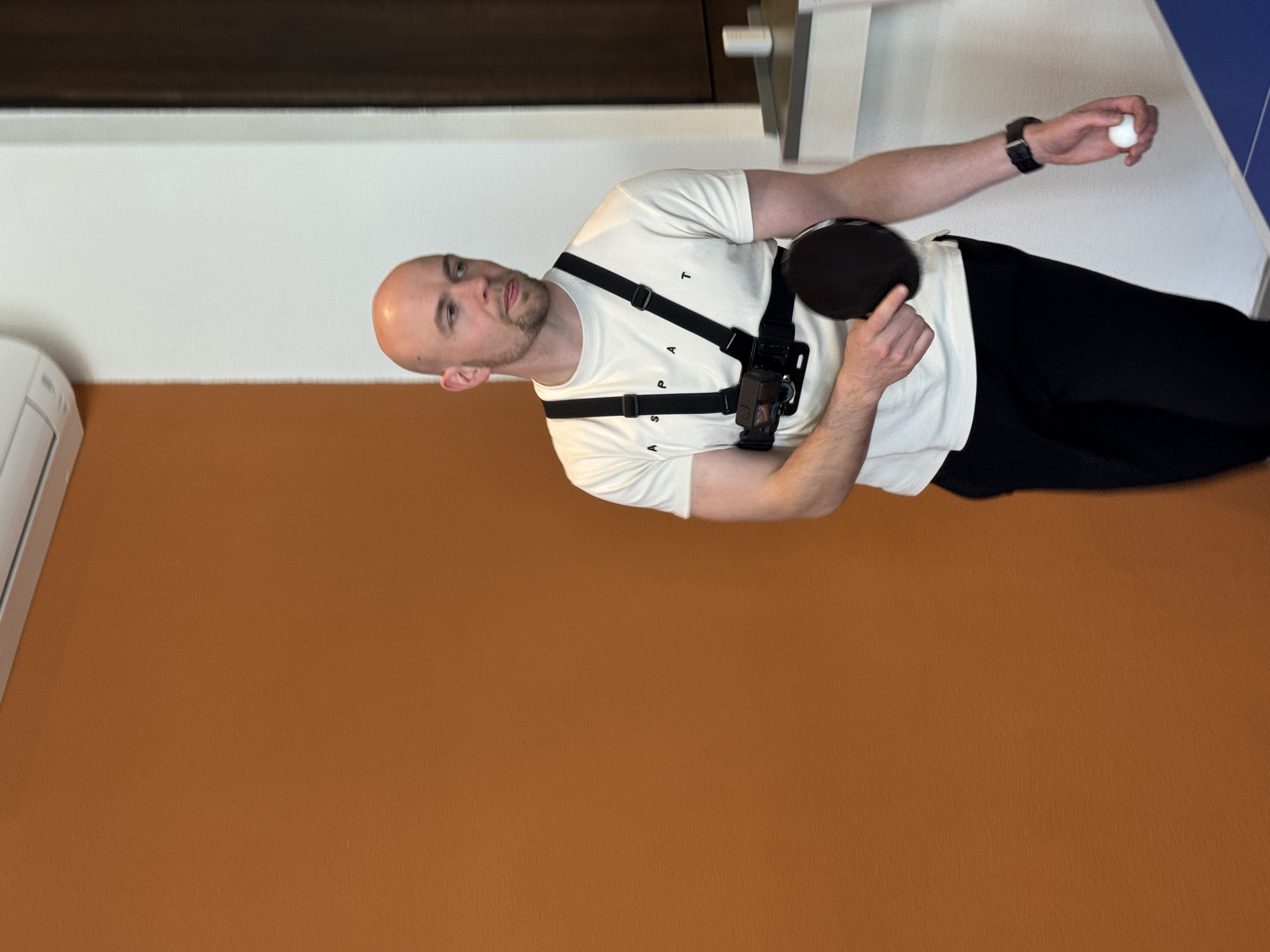 A bald man holds a ping pong ball and paddle, wearing a white vest.