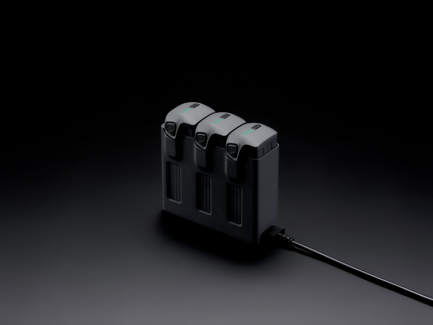 Tre batterie grigie per droni in carica in un hub di ricarica nero collegato a un cavo.