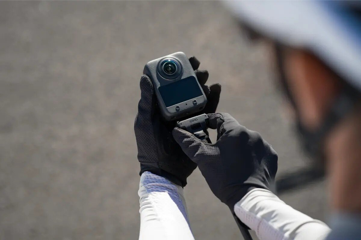 Hände in schwarzen Handschuhen halten eine graue Kamera mit Bildschirm und Linse. Draußen.