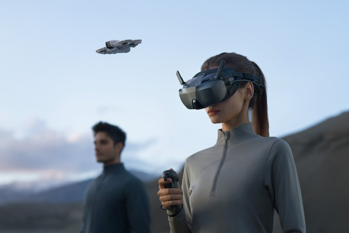 Donna con occhiali VR tiene controller. Uomo e drone sullo sfondo, esterno.