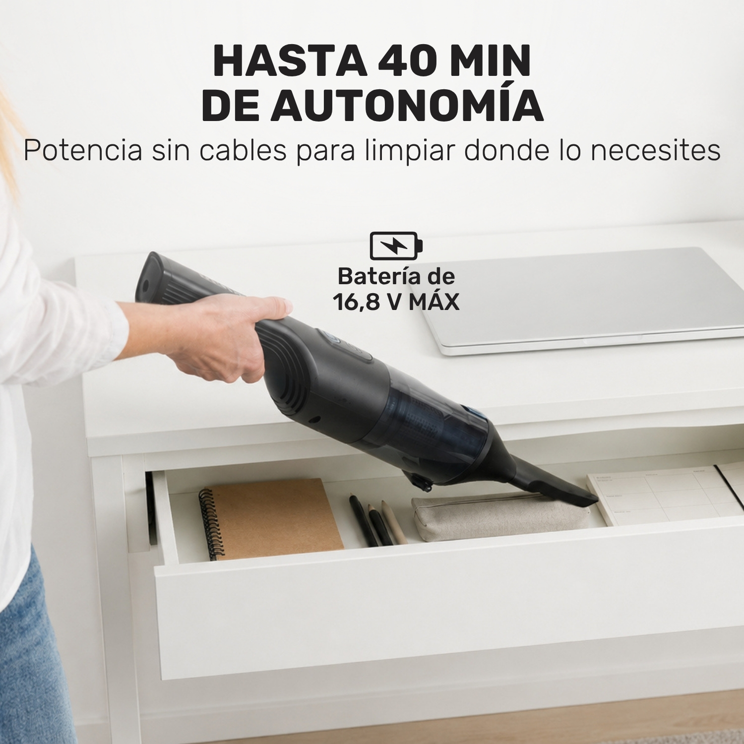 Una mano sostiene una aspiradora negra de mano que entra en un cajón con un cuaderno y bolígrafos.