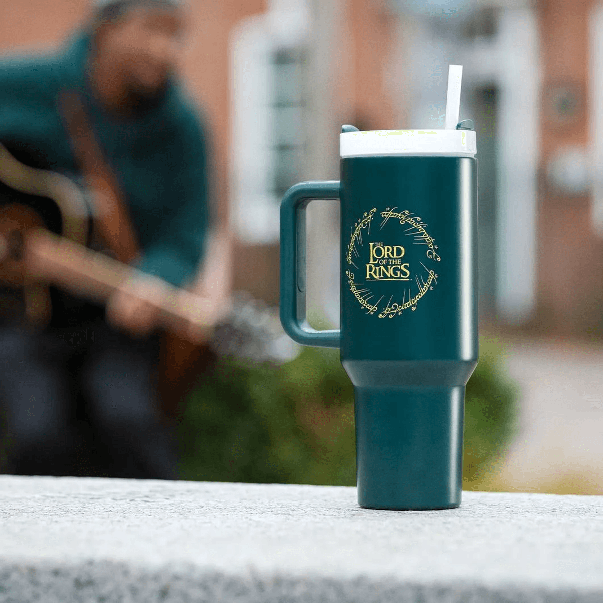 Una taza verde azulado de El Señor de los Anillos con pajita, sobre una repisa de piedra. Un guitarrista desenfocado al fondo.