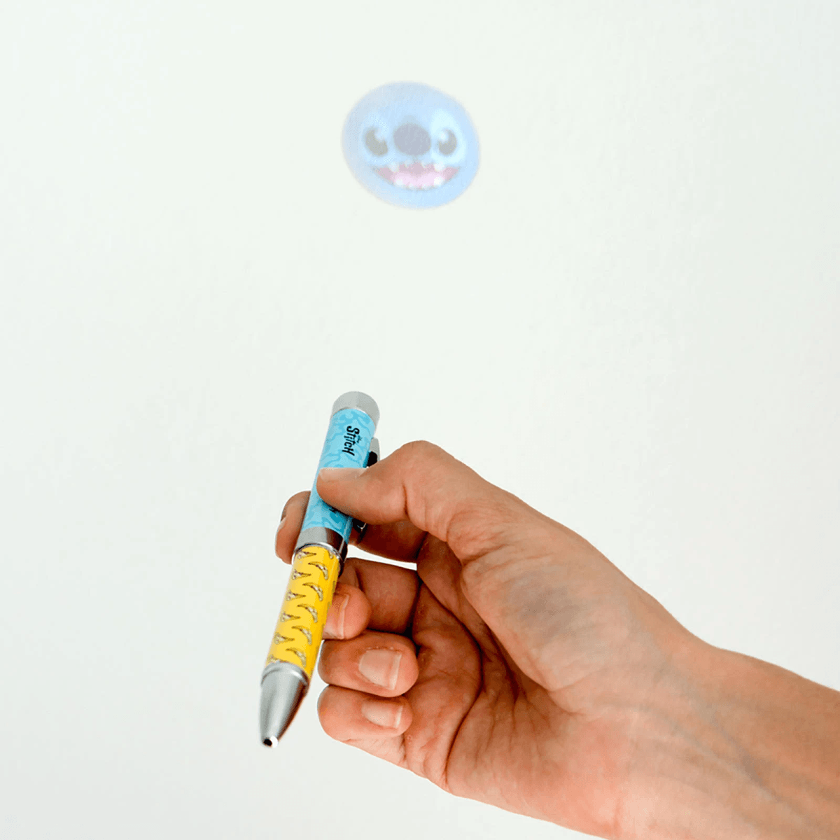 Una mano sostiene un bolígrafo azul y amarillo que proyecta una imagen de la cara azul de Stitch.