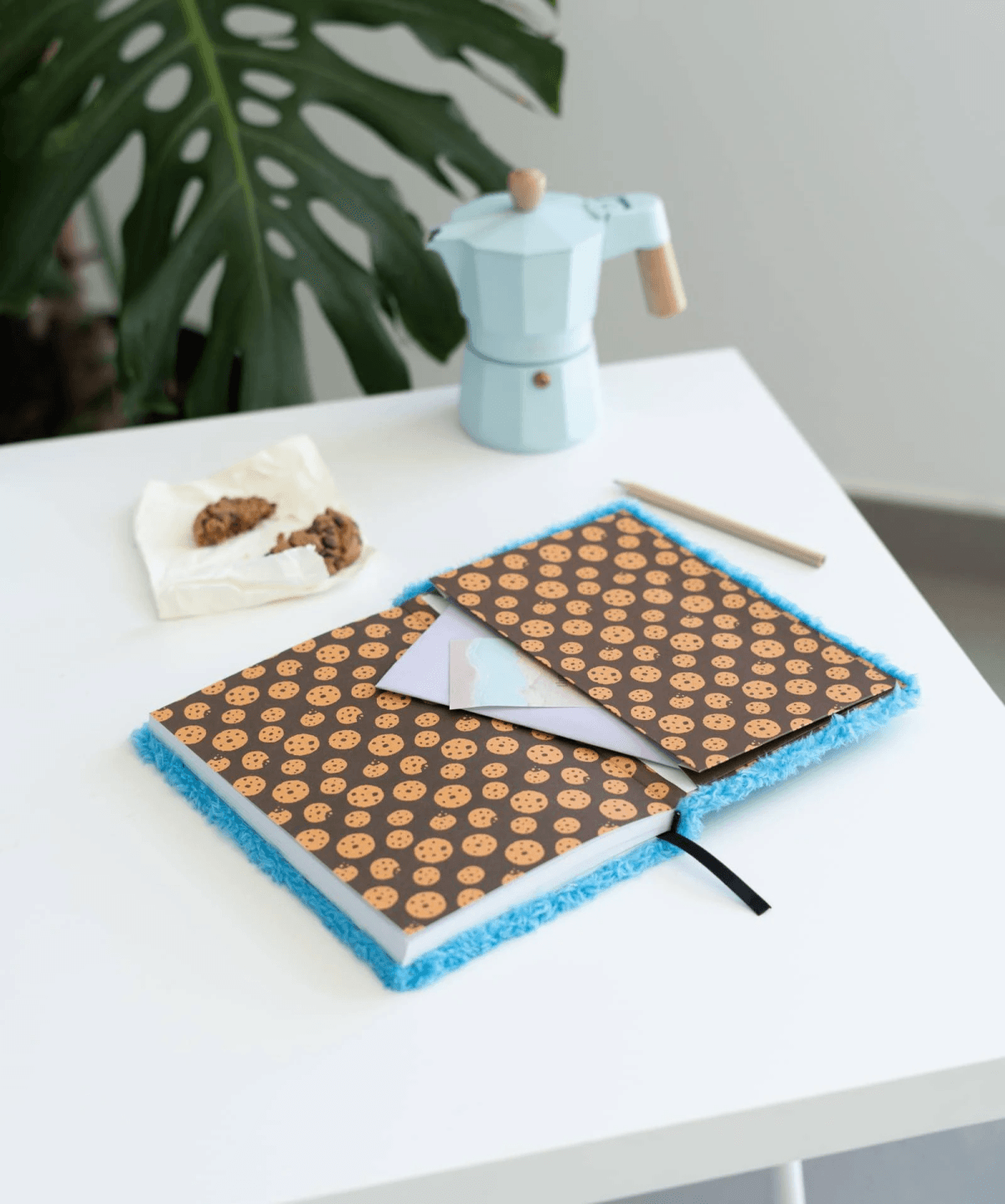 Un cuaderno abierto con estampado de galletas y borde azul, junto a una cafetera moka.